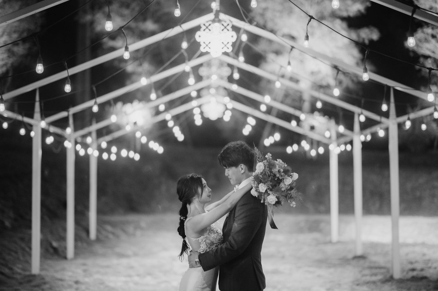 Romantic couple shares first dance under glowing string lights in black and white wedding photograph.