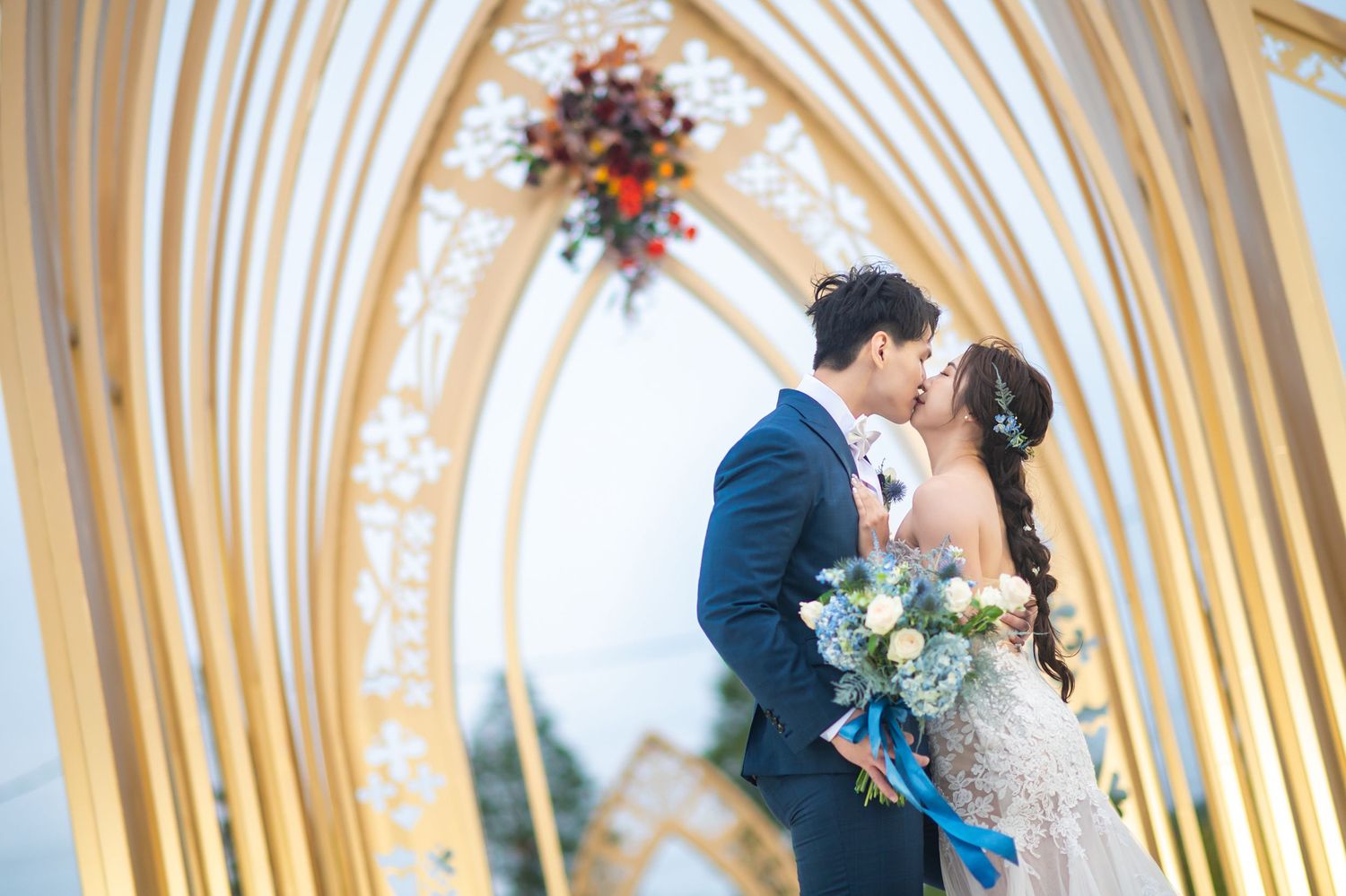 A romantic moment between newlyweds under a golden archway decorated with floral arrangements at sunset.