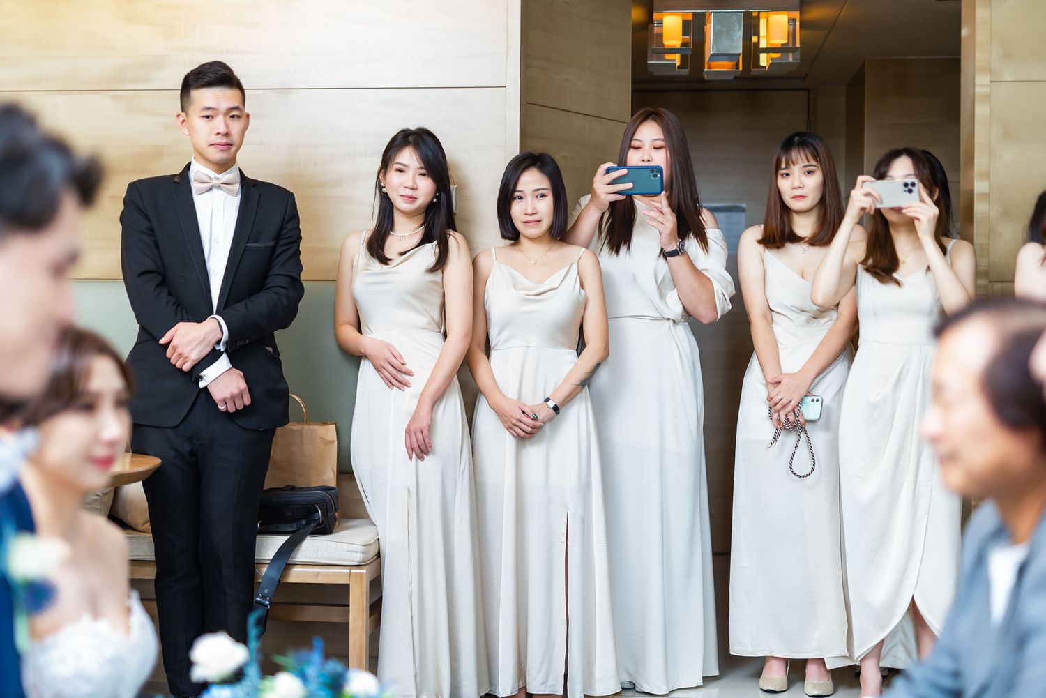 A formal wedding party wearing matching white dresses and suits gathers for photos in an elegant indoor setting.
