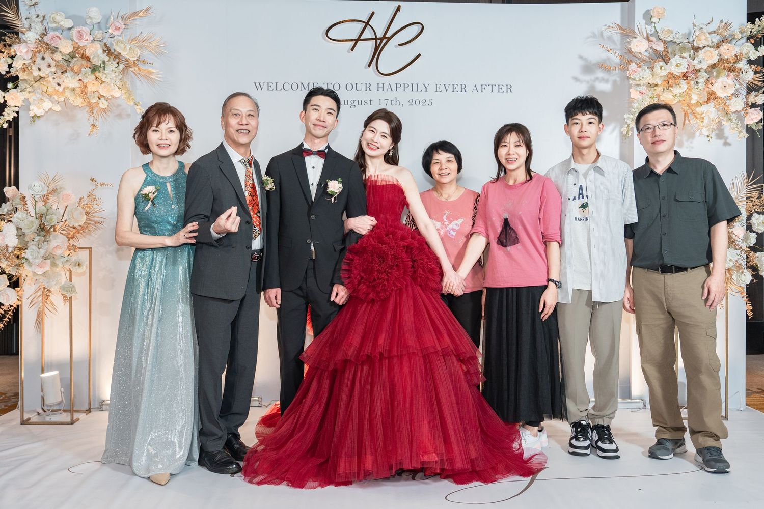 Group photo at wedding reception with bridesmaids in blue dresses and bride in red tulle gown with cherry blossoms.