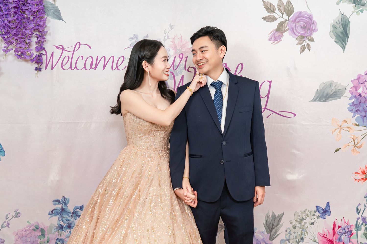 A couple in formal attire share a romantic moment against a floral backdrop during their wedding photoshoot.