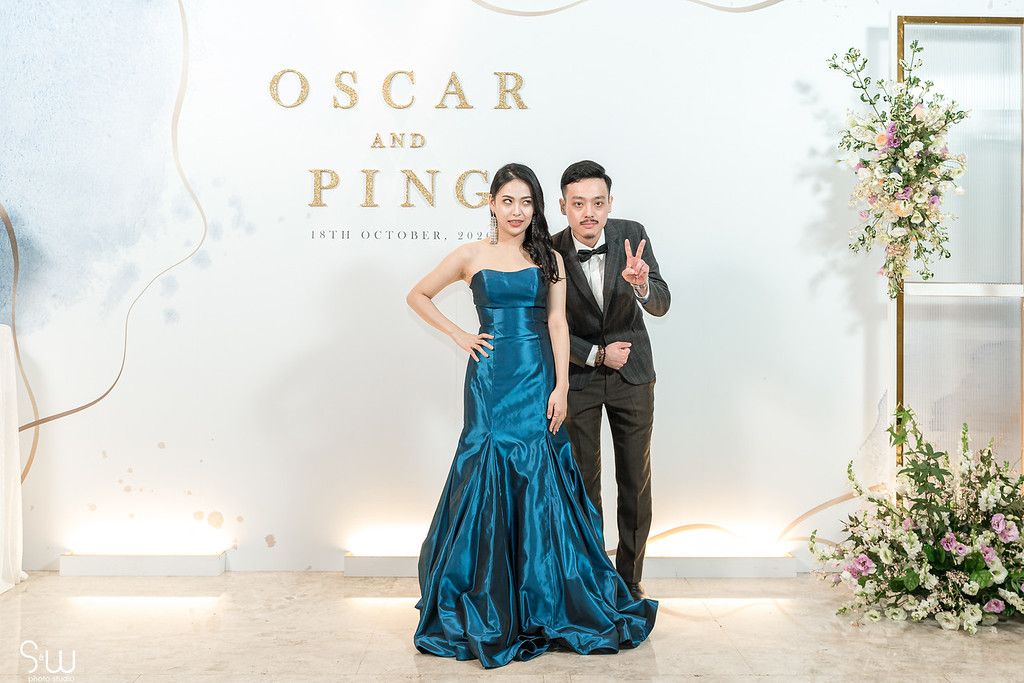 A couple strikes a playful pose in formal attire against a white backdrop with gold lettering.