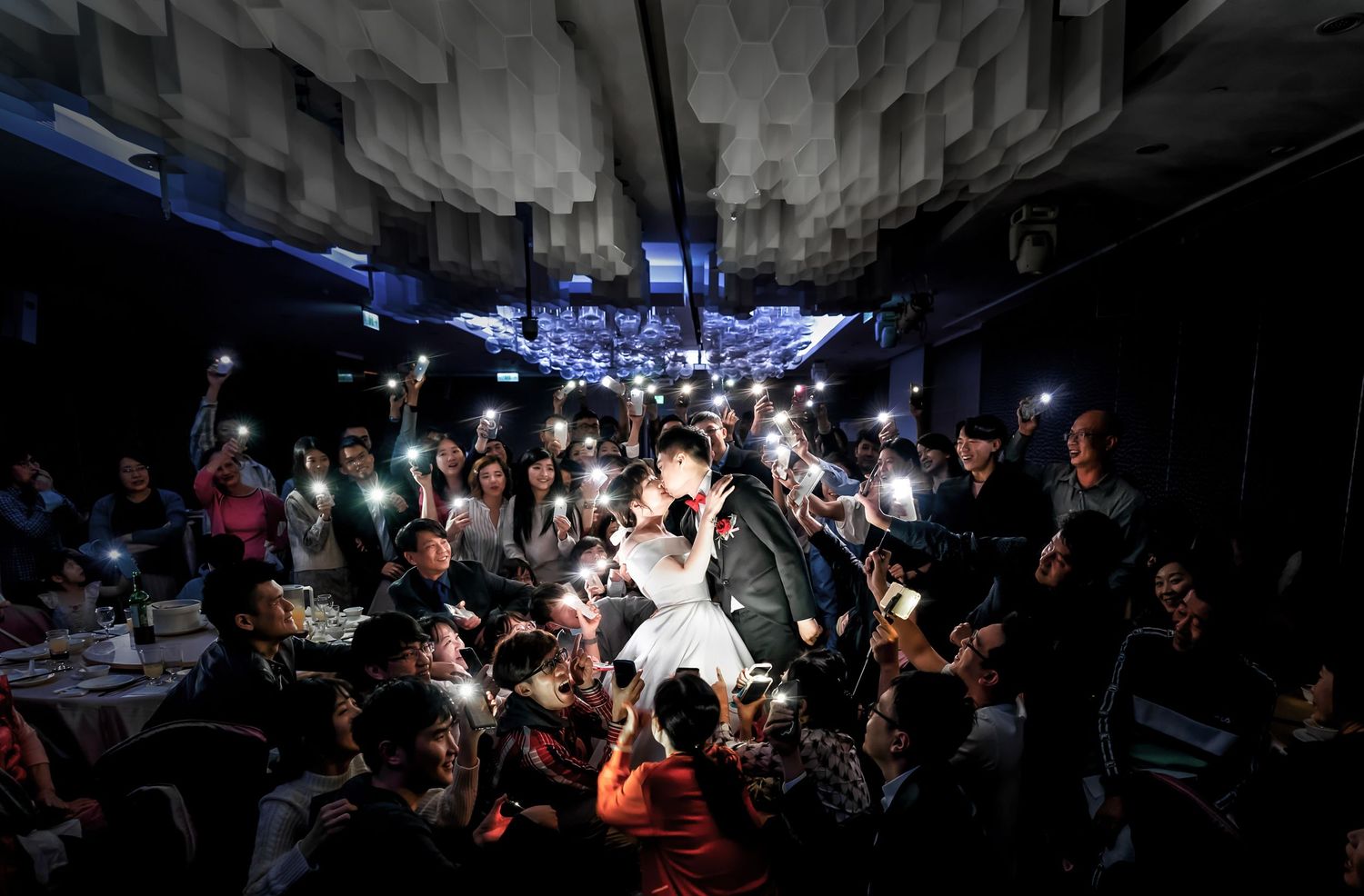 Guests surround the wedding couple in a dark venue while holding up phone lights creating a dramatic effect.