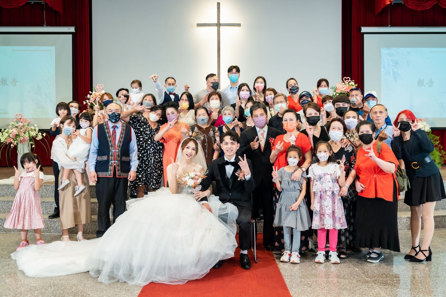 Large group wedding portrait with guests gathered on red carpet steps in church setting.