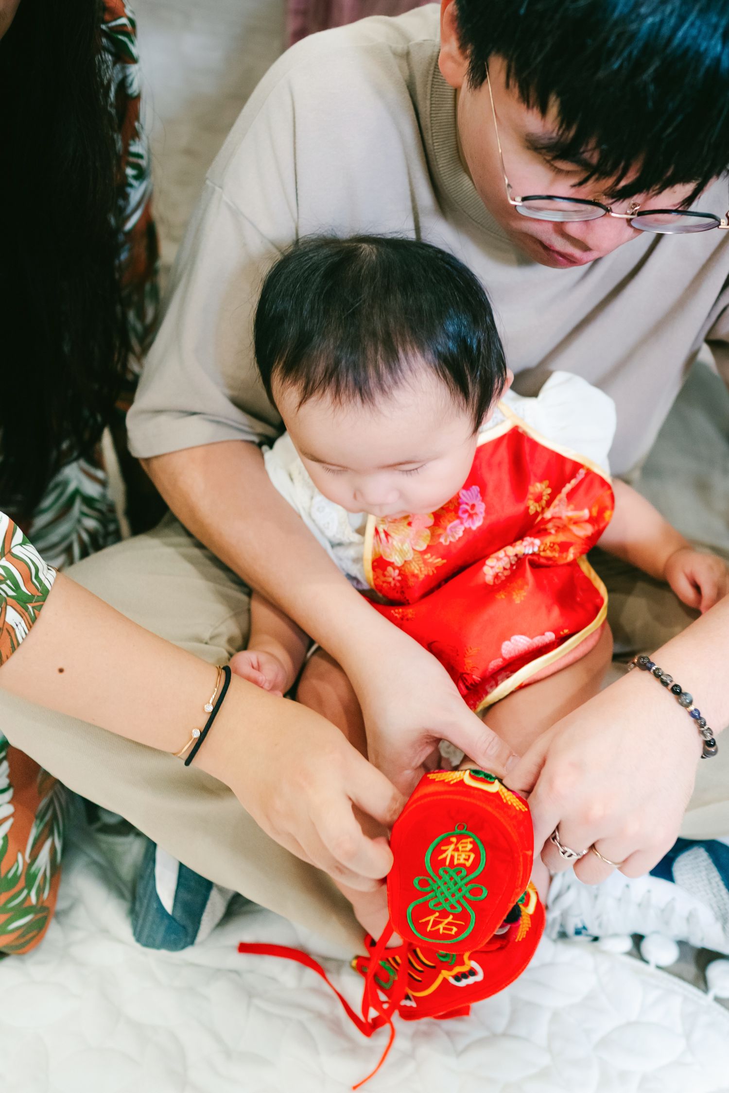 A baby wearing traditional red Chinese New Year clothing sits with a parent holding a red lucky envelope decoration.