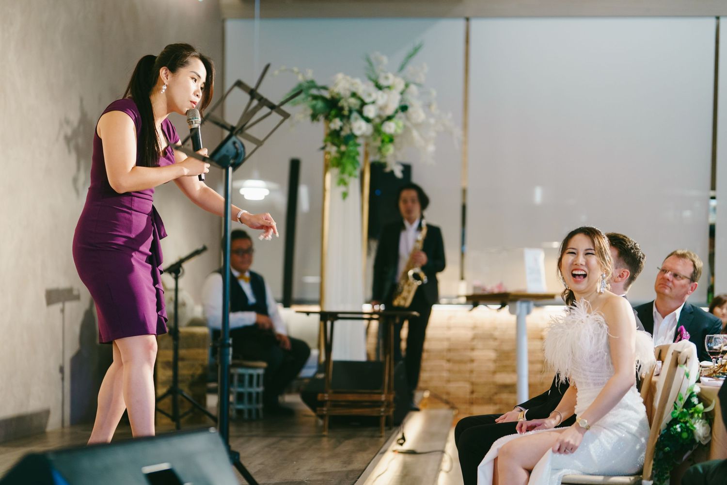 People gather for an indoor event with someone in a purple dress speaking while others sit and watch with laughter.