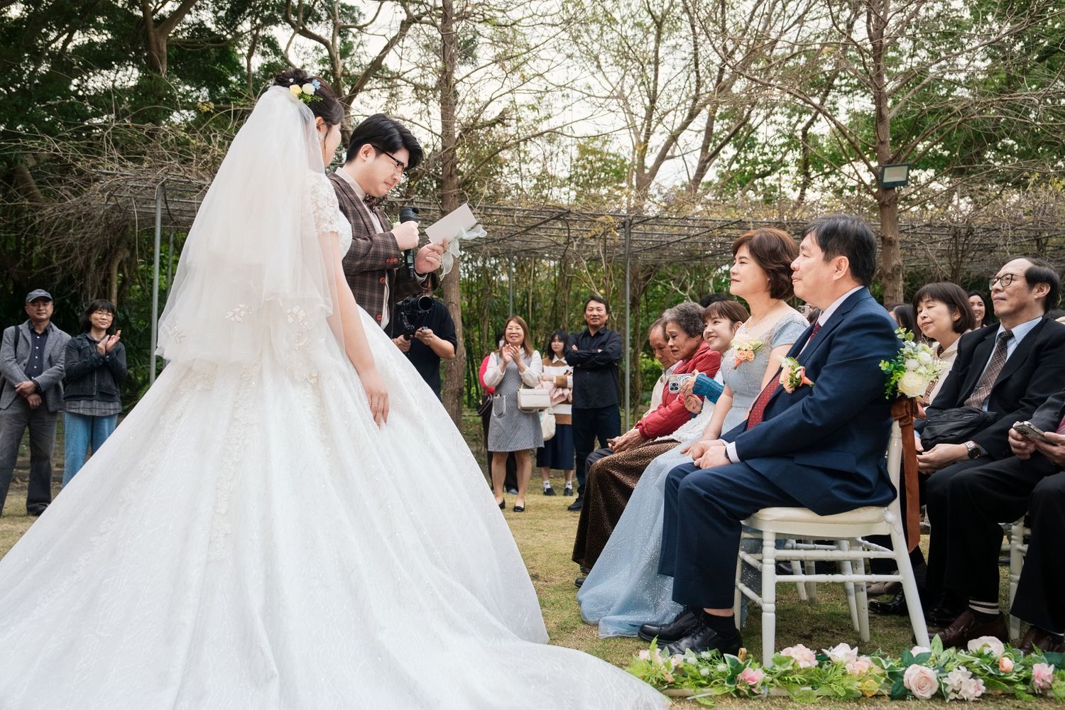 Wedding ceremony guests gather outdoors in a garden setting as a bride in white dress walks down the aisle.