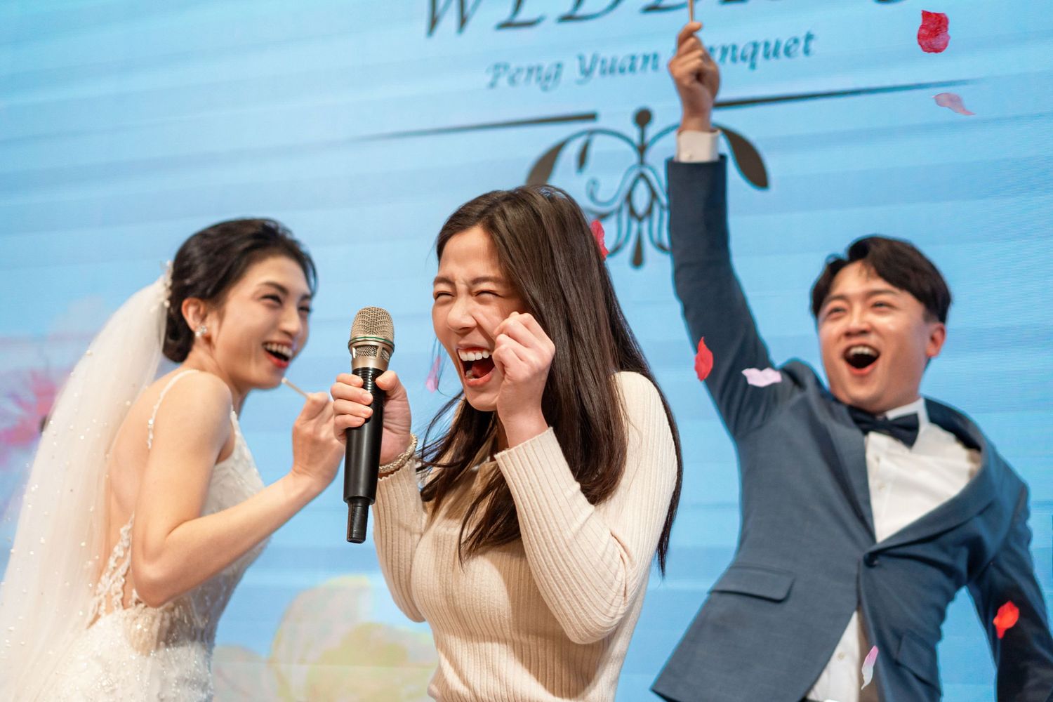 Joyful celebration at a wedding reception while guests sing karaoke with microphones against a blue backdrop.