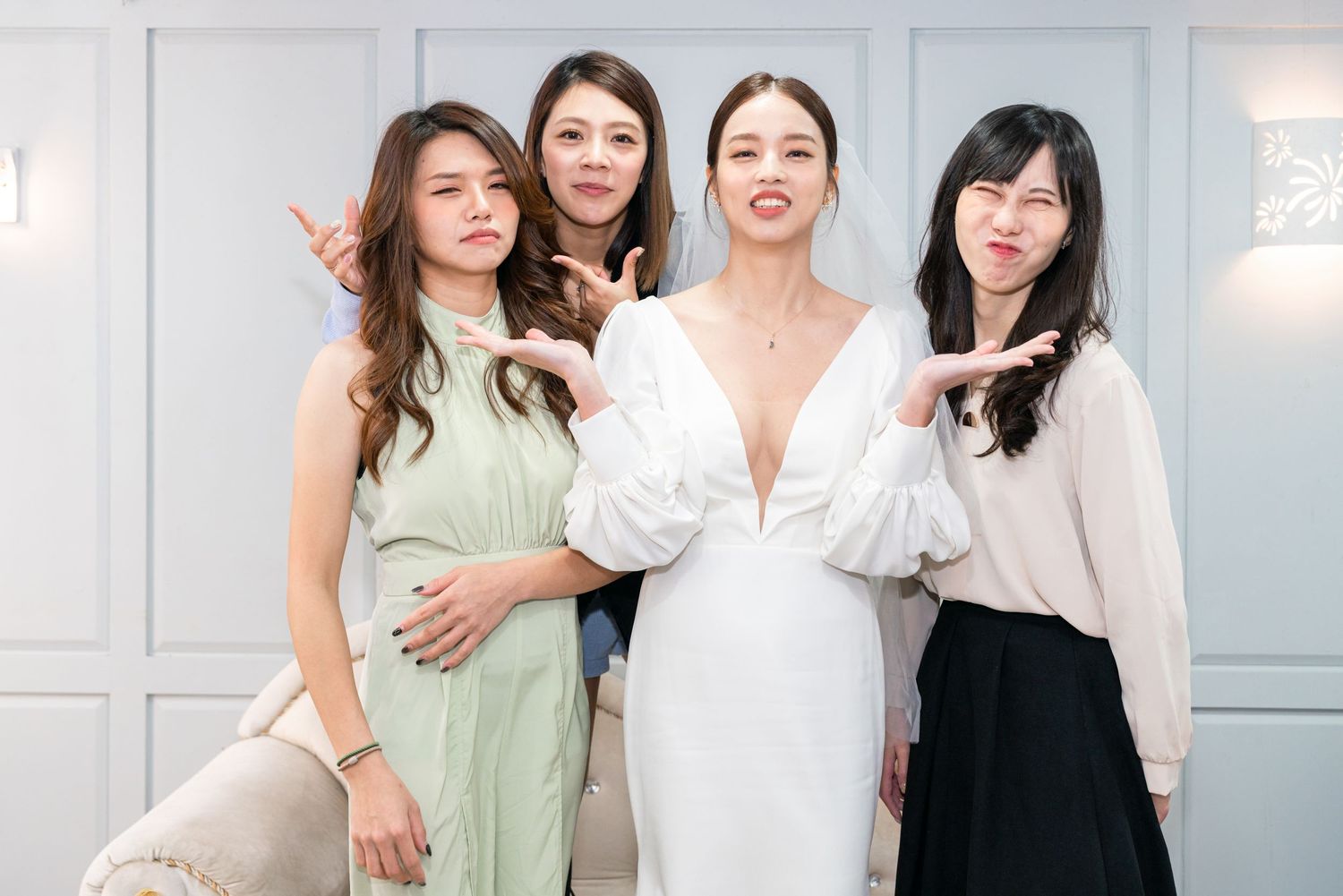 Four friends in elegant attire pose together at a wedding celebration with white decorative wall panels in background.