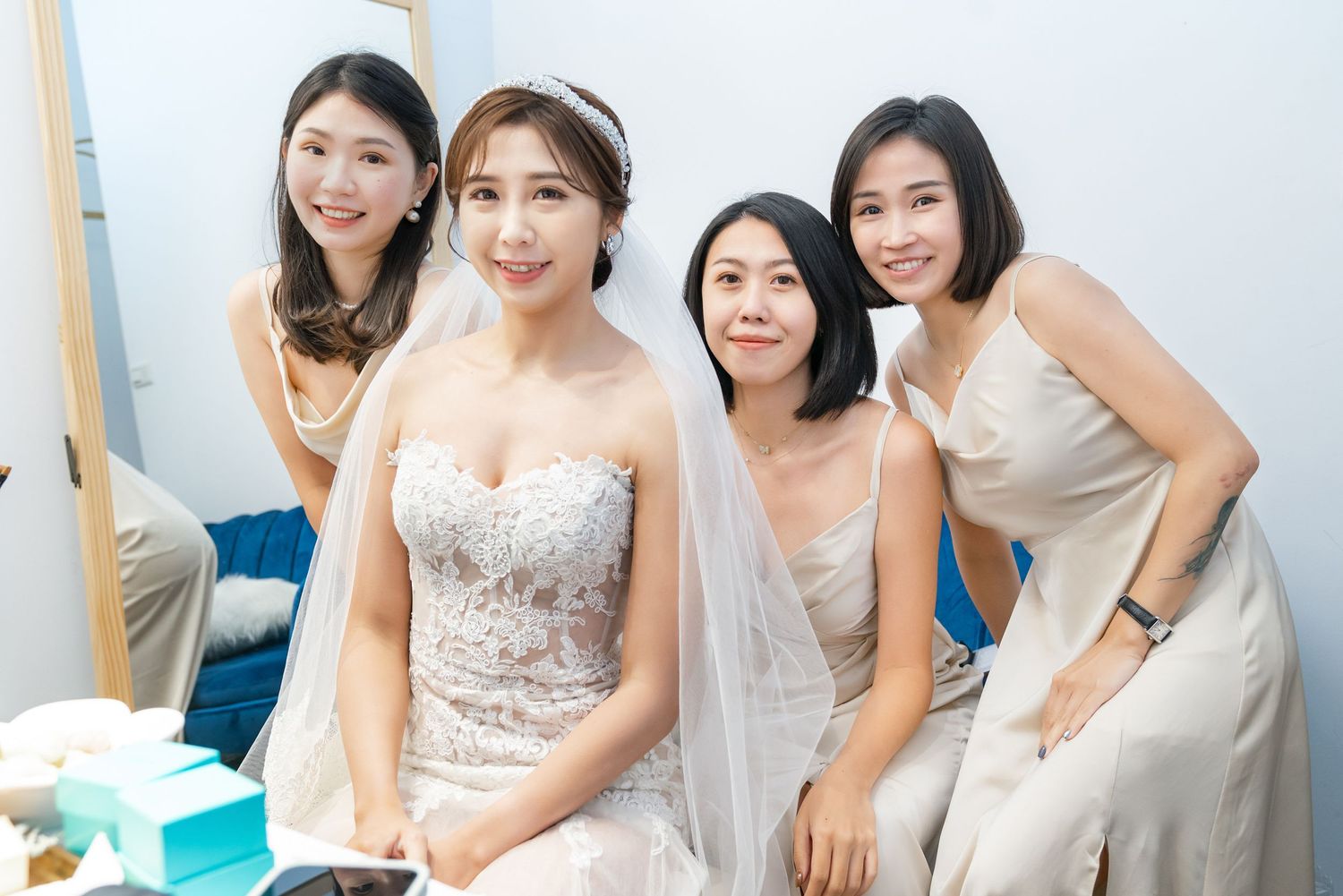 A group smiles together in a bridal room as they gather around a person wearing an elegant strapless white wedding gown.
