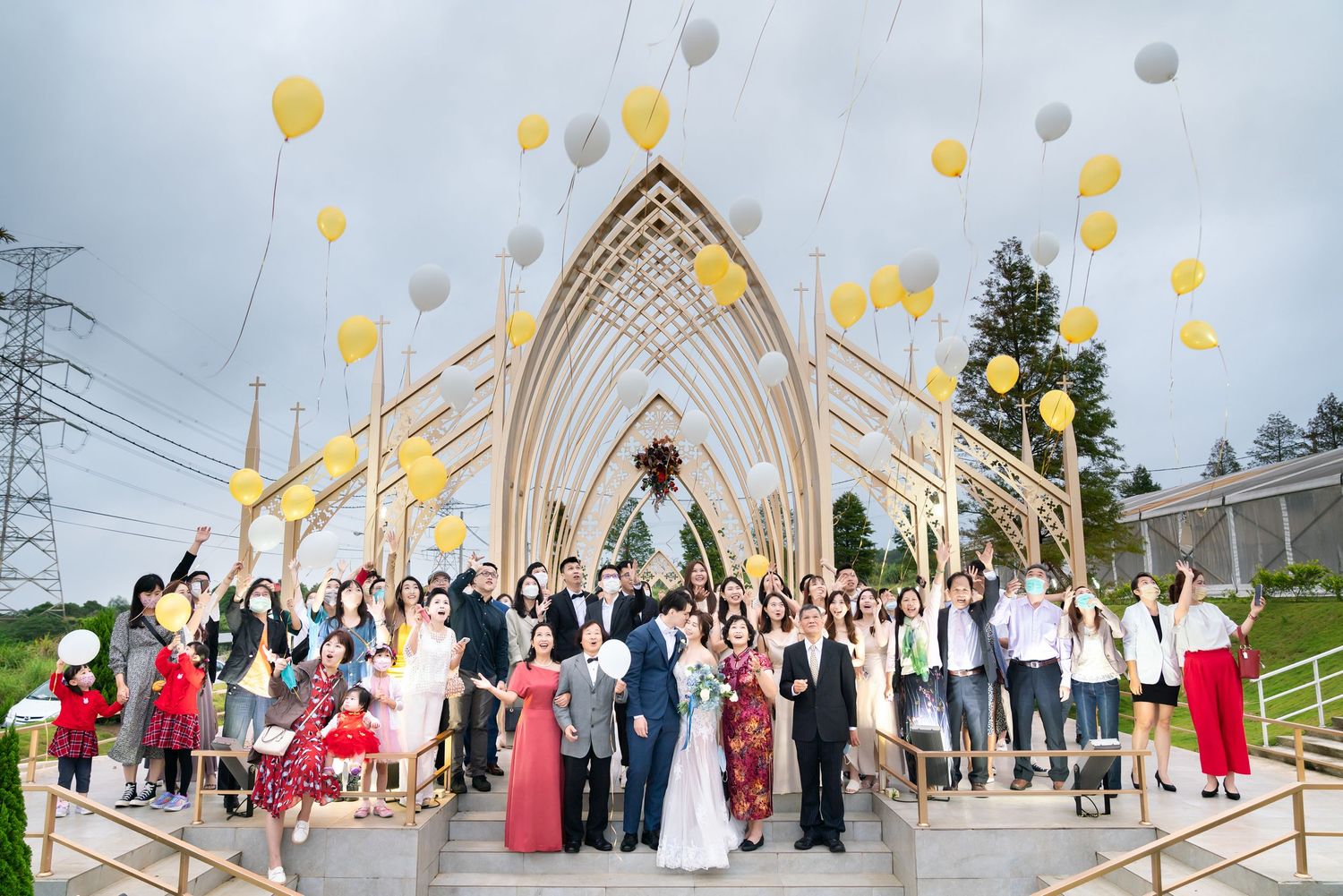 A large group of wedding guests release yellow and white balloons outside a modern chapel with an arched entrance.