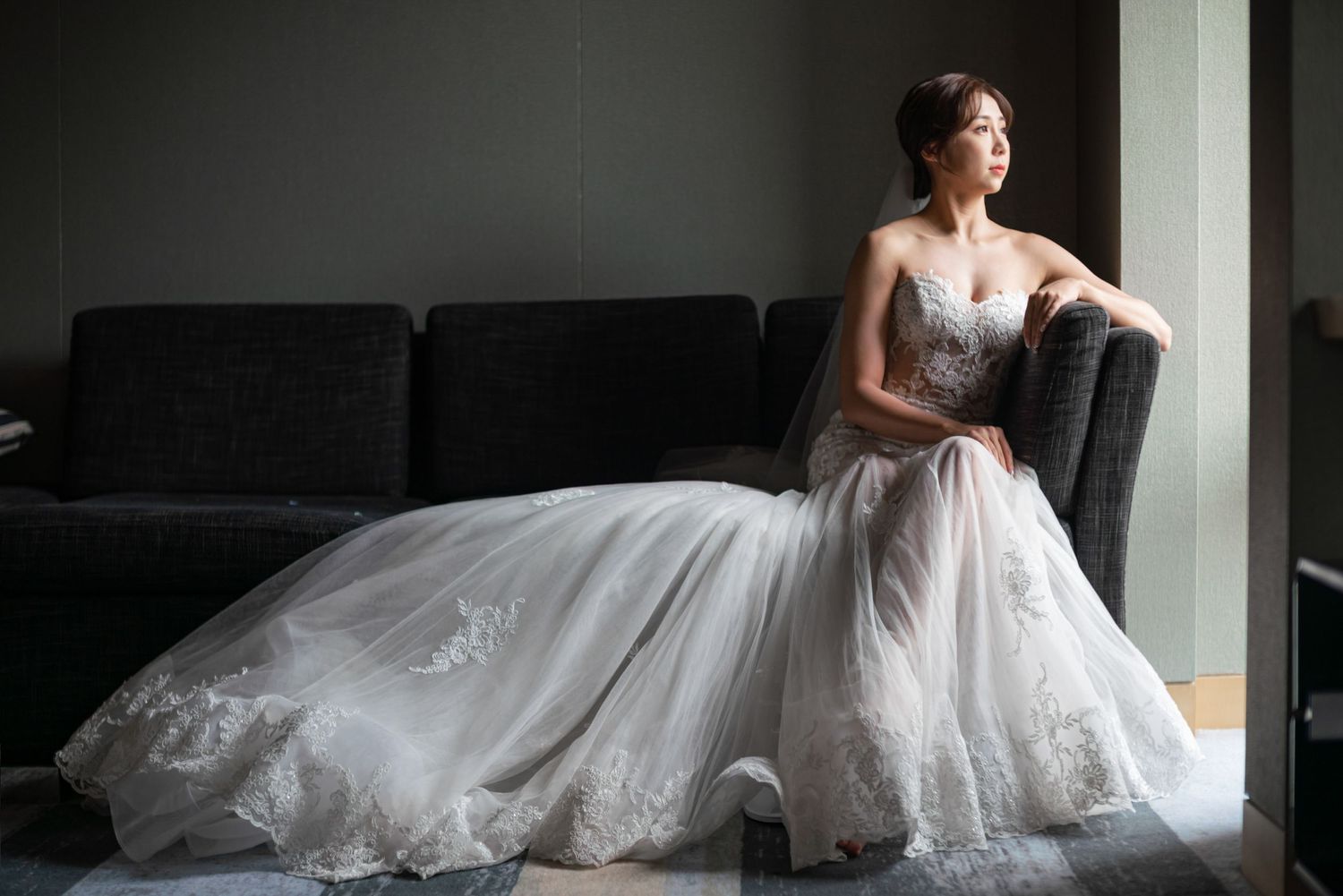 A bride in an elegant white lace gown sits on a dark couch near a window in dramatic natural lighting.