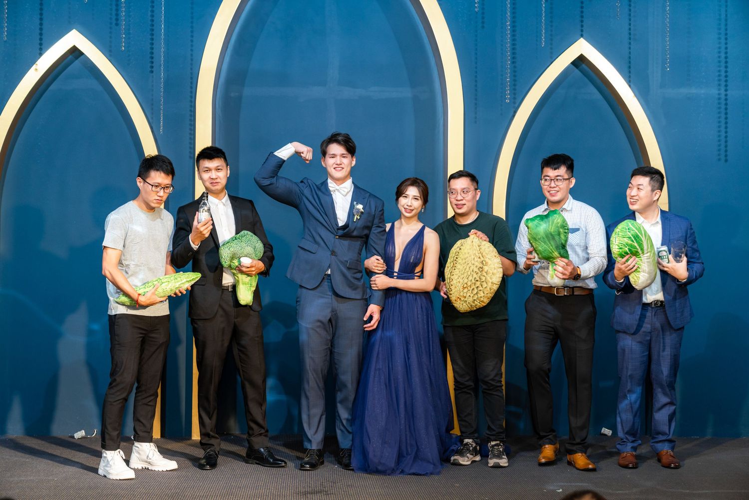 A group poses with tropical fruits against a blue wall wearing formal and casual attire.