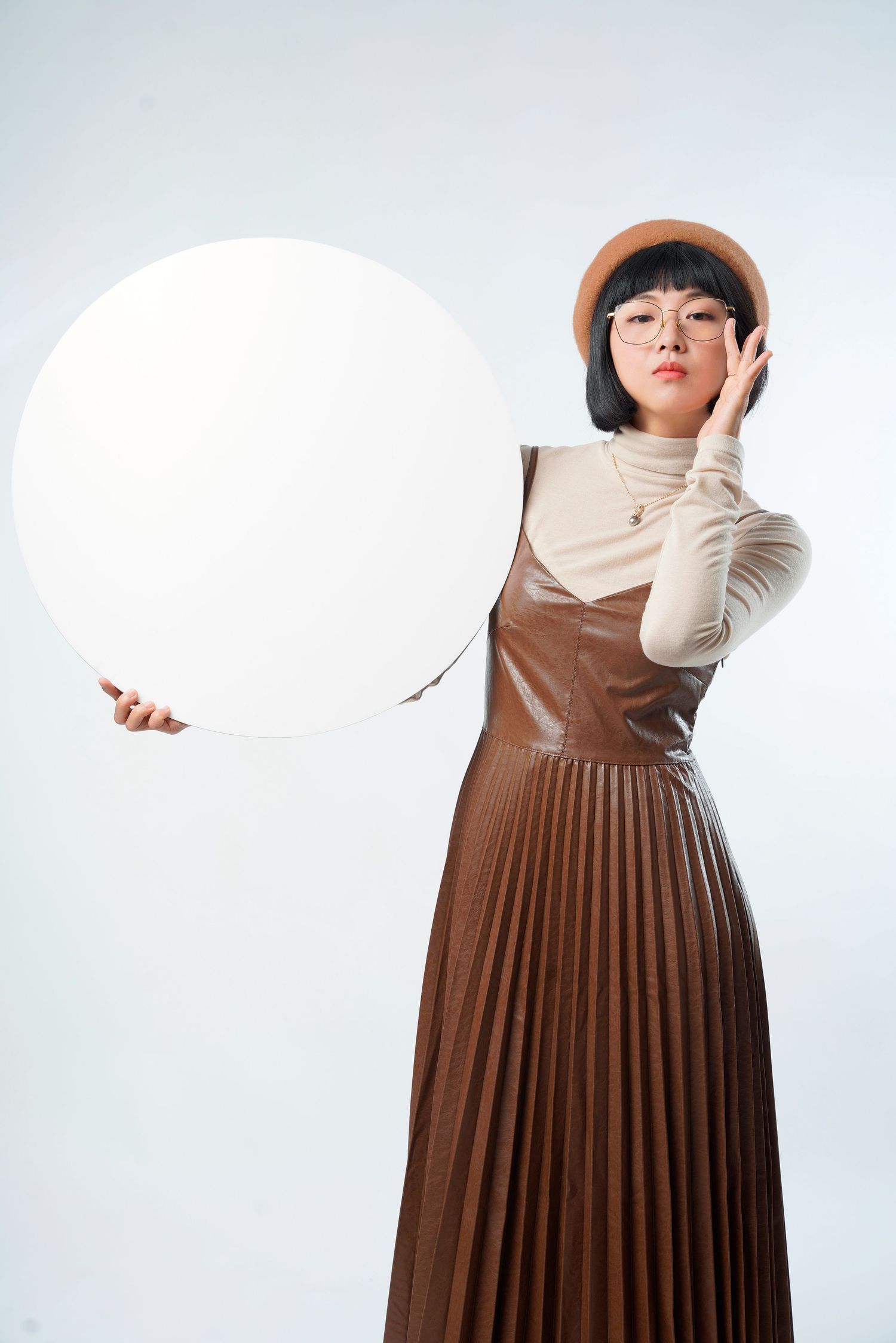 Fashion model in brown pleated maxi dress and white turtleneck holds large white sphere against white backdrop.