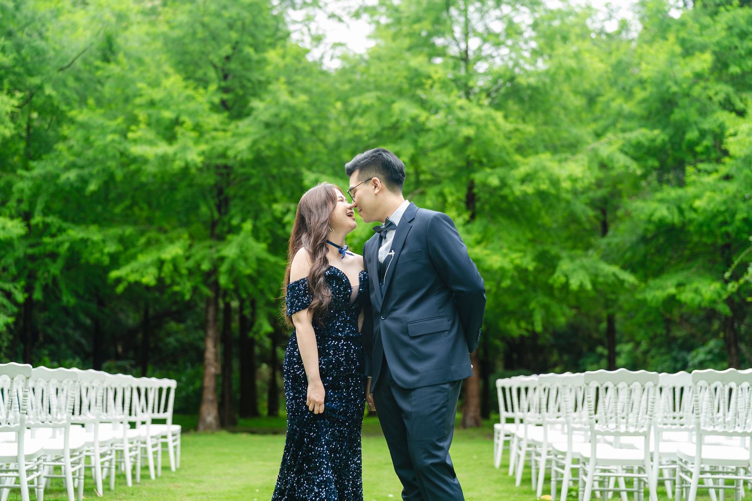 Couple sharing a tender moment on a lush green lawn with white ceremony chairs arranged behind them.