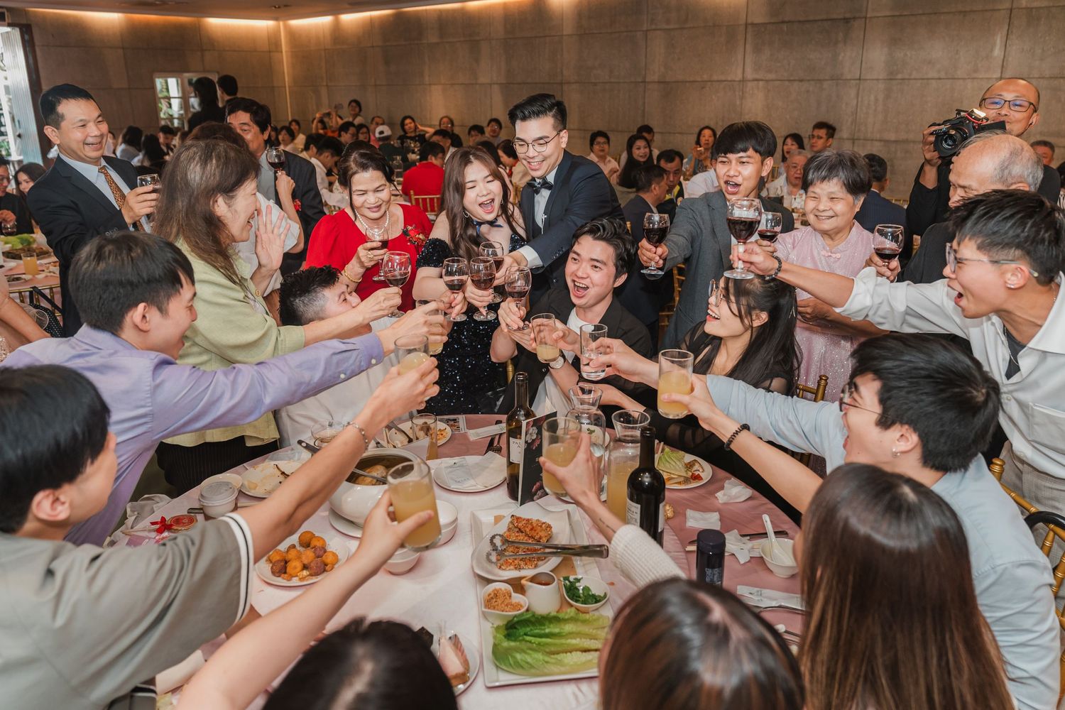 A large group toasts with drinks at a crowded restaurant table during a lively celebration gathering.