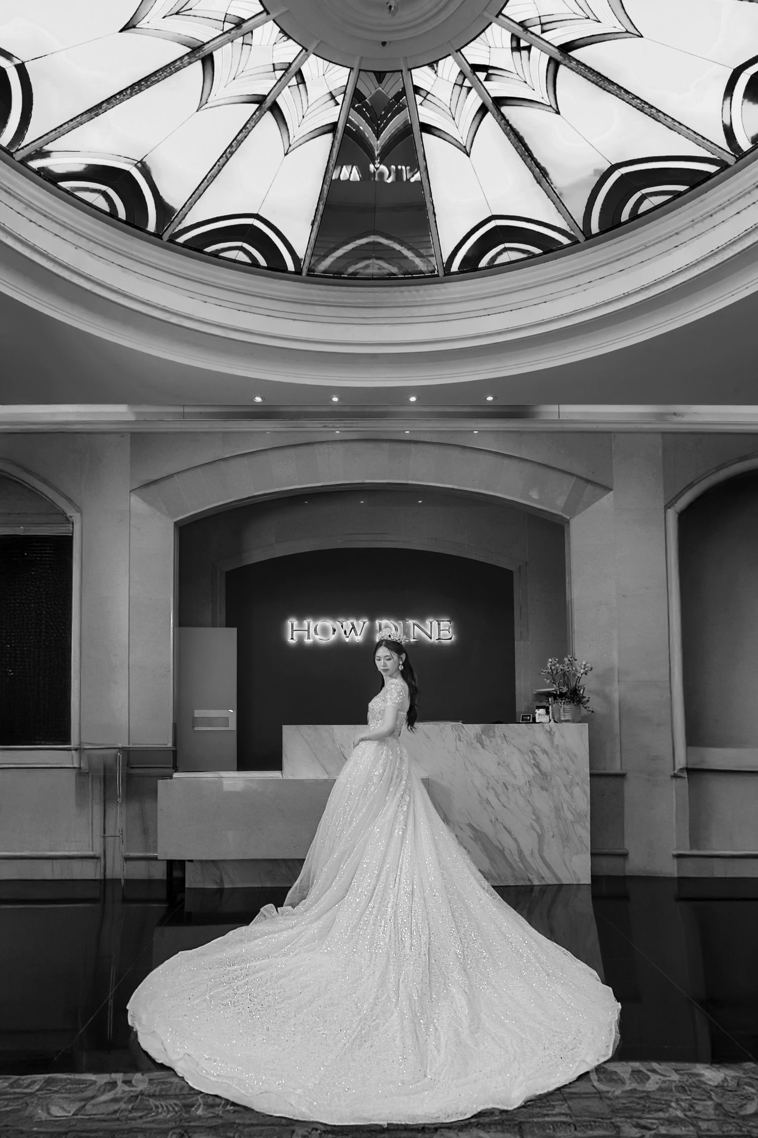 A bride in an elegant white gown with a dramatic cathedral-length train stands in front of a hotel reception desk.