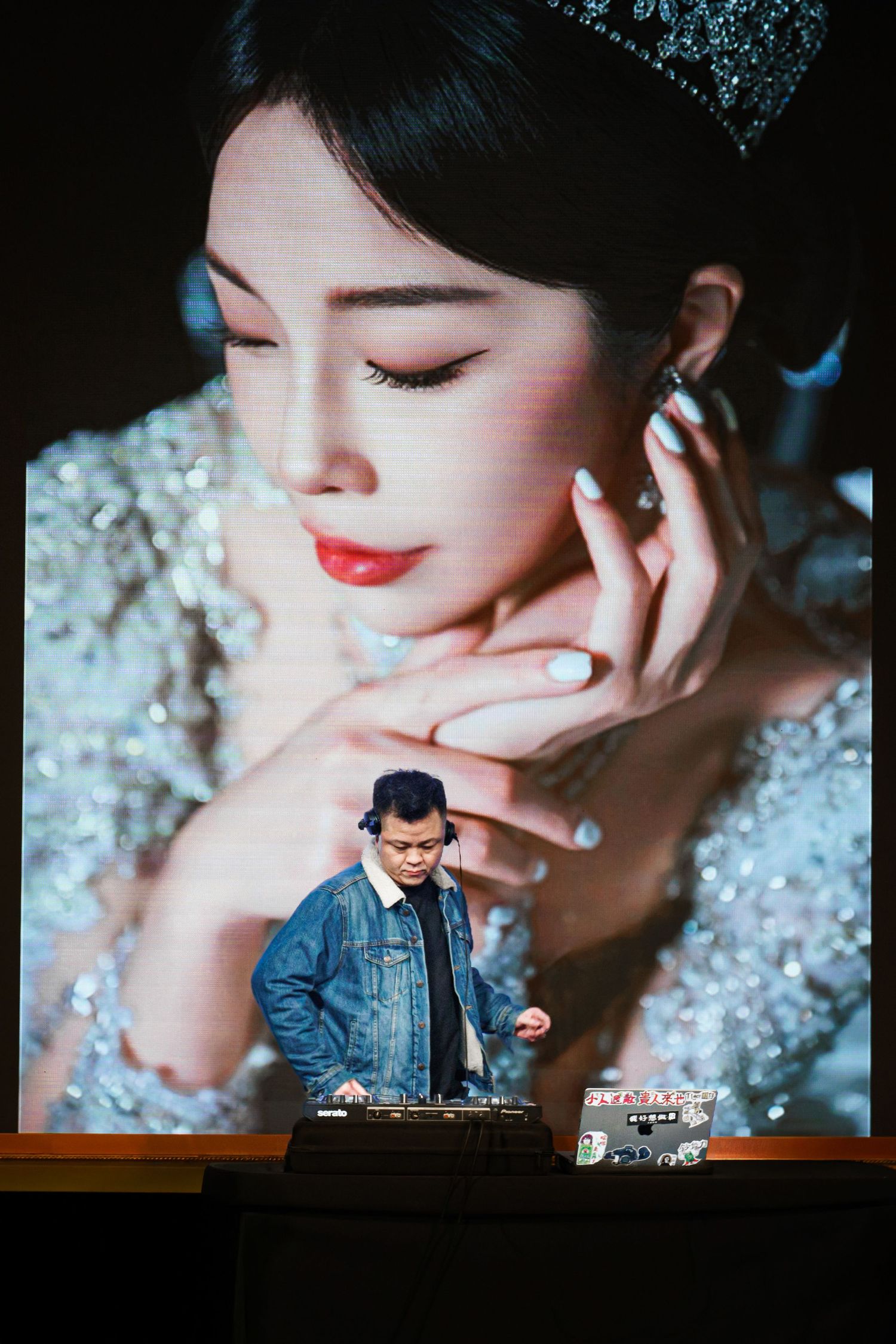 DJ performs at event with large artistic portrait projections displayed behind mixing equipment on stage.