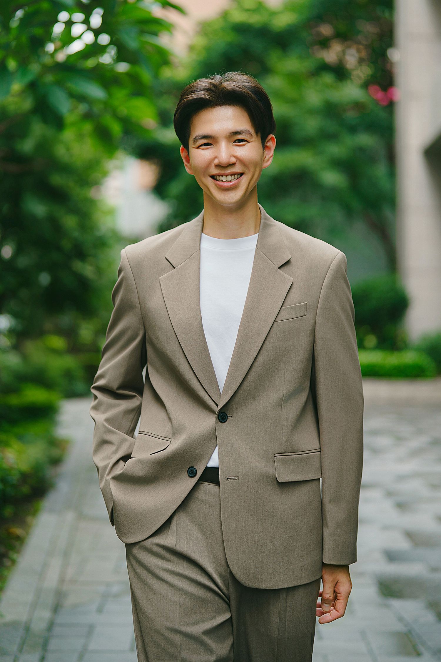 A person wearing a beige suit and white t-shirt smiles while standing on a stone pathway surrounded by greenery.
