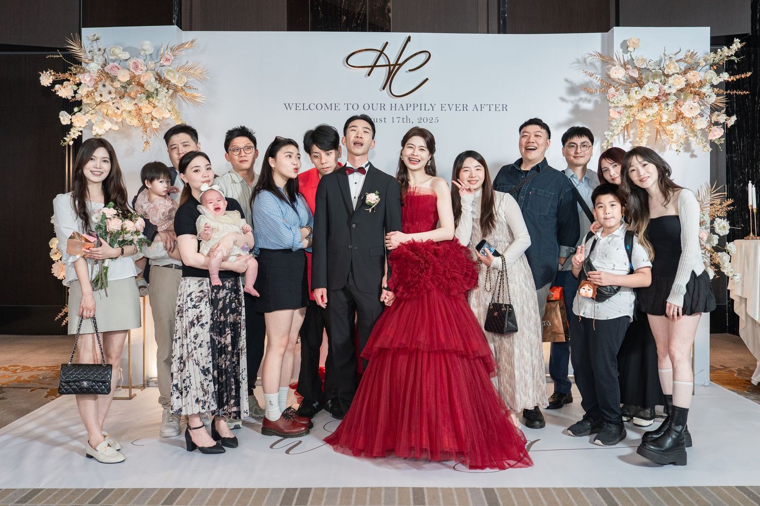 Group photo at an elegant wedding reception with a bride in a red layered gown in front of a decorated backdrop.