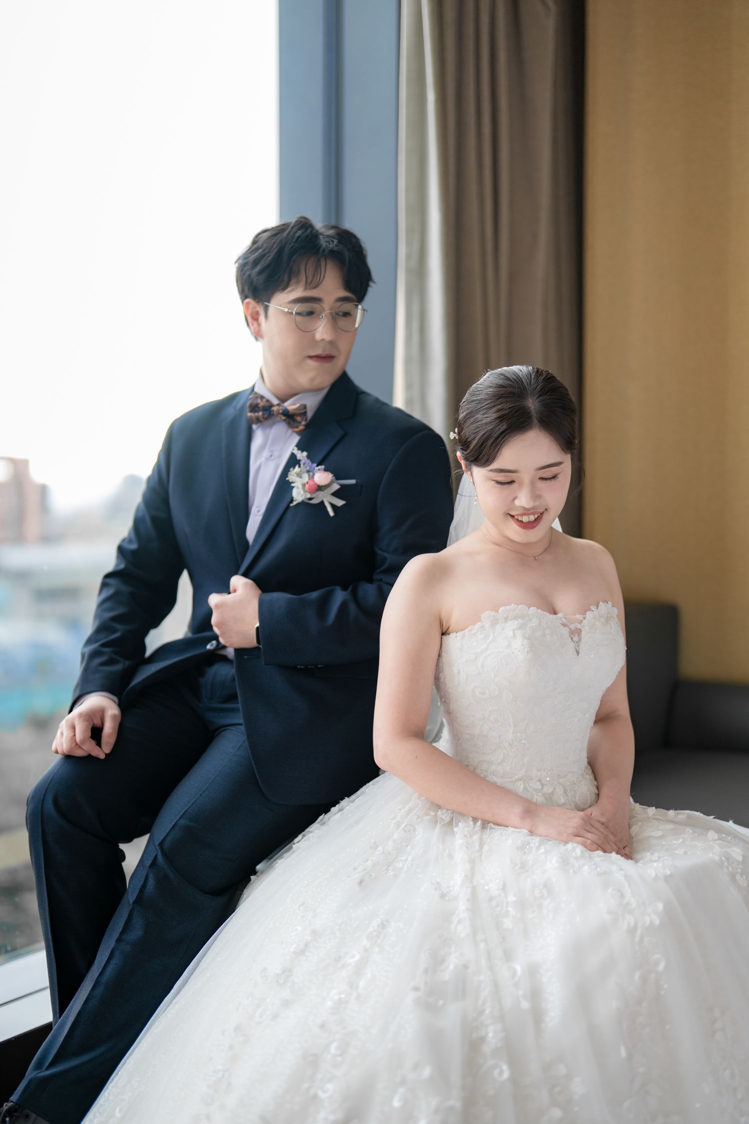 A bride in a white strapless gown and groom in black suit share tender moments in a sunlit room.