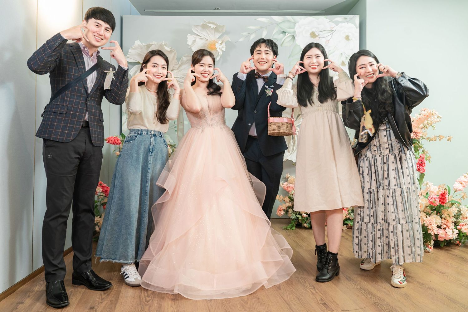 A group of wedding guests pose together making heart gestures in front of a floral backdrop.