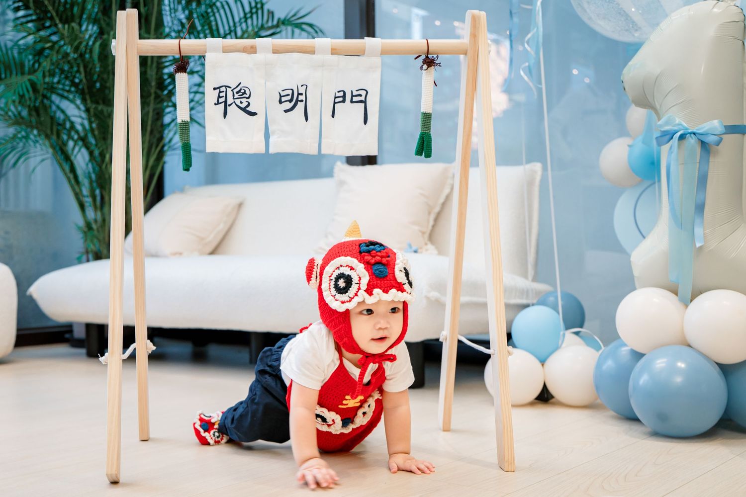 Baby in red Chinese zodiac costume crawls under wooden frame with decorative balloons and Asian calligraphy banner.