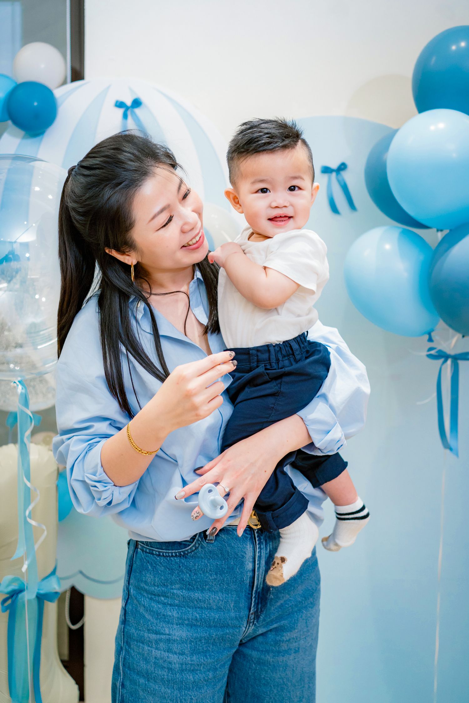A joyful celebration with blue and white balloons decorating the background as parent and child share a happy moment together.