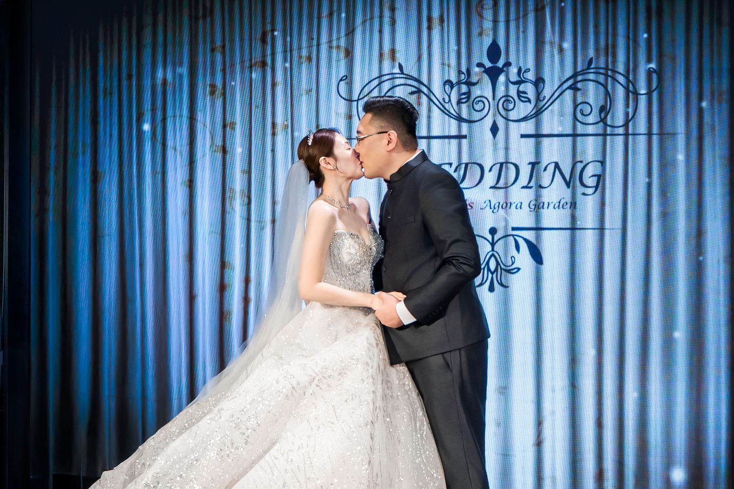 A couple shares a romantic kiss at their wedding with a blue curtain backdrop and elegant decor.