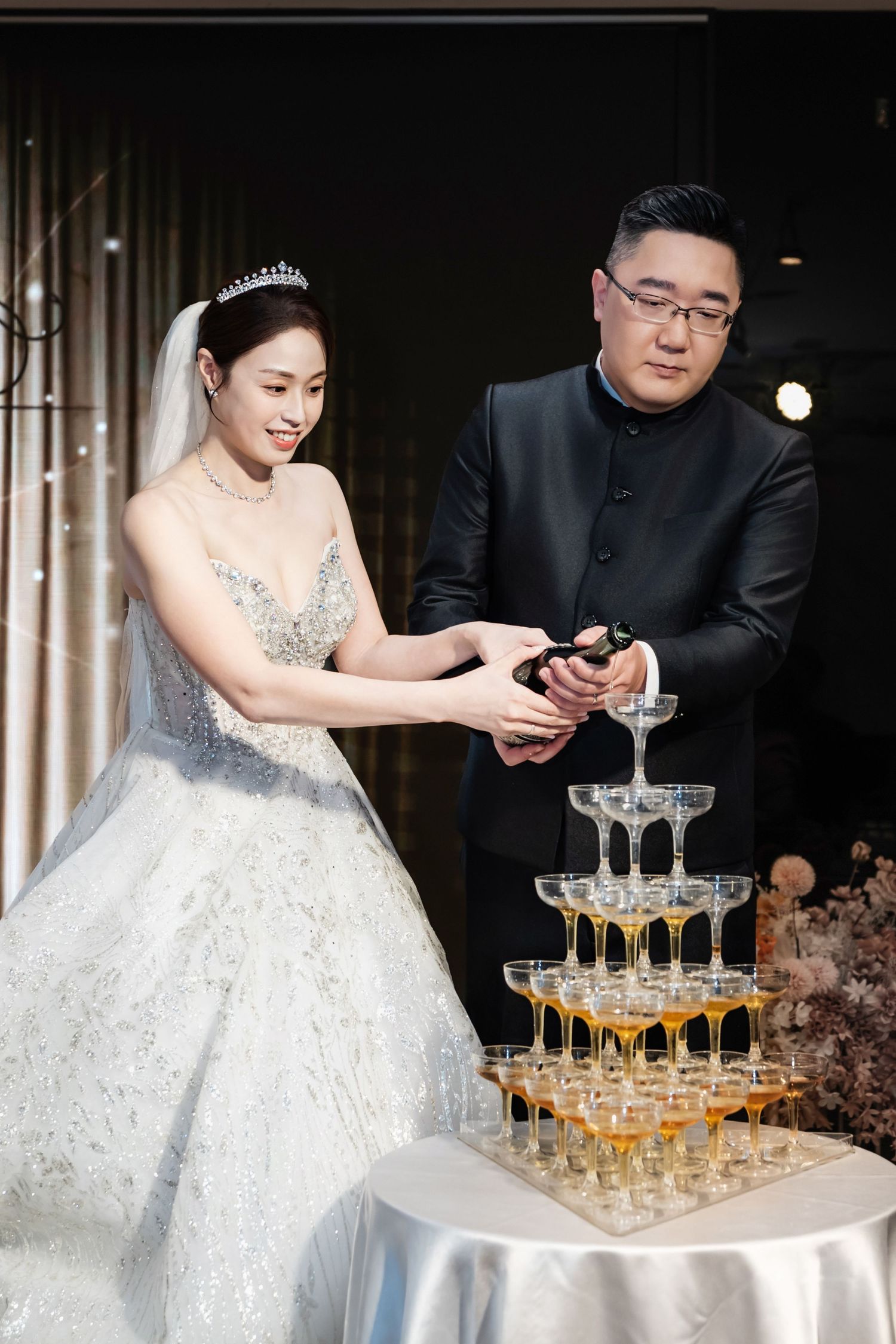 A couple pours champagne into a tower of crystal glasses during their wedding celebration.