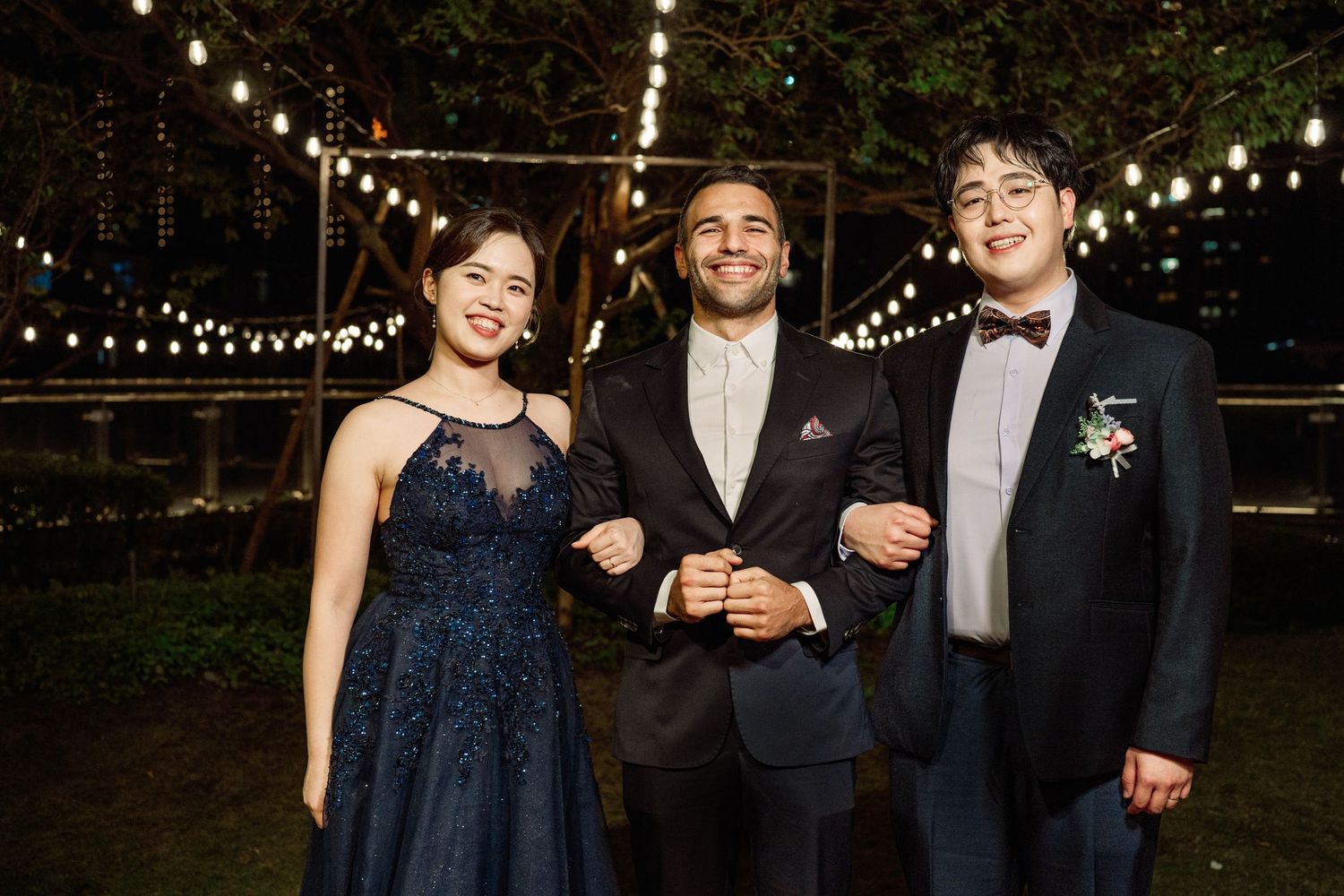 Three people in formal attire smile together at an outdoor evening event with string lights overhead.