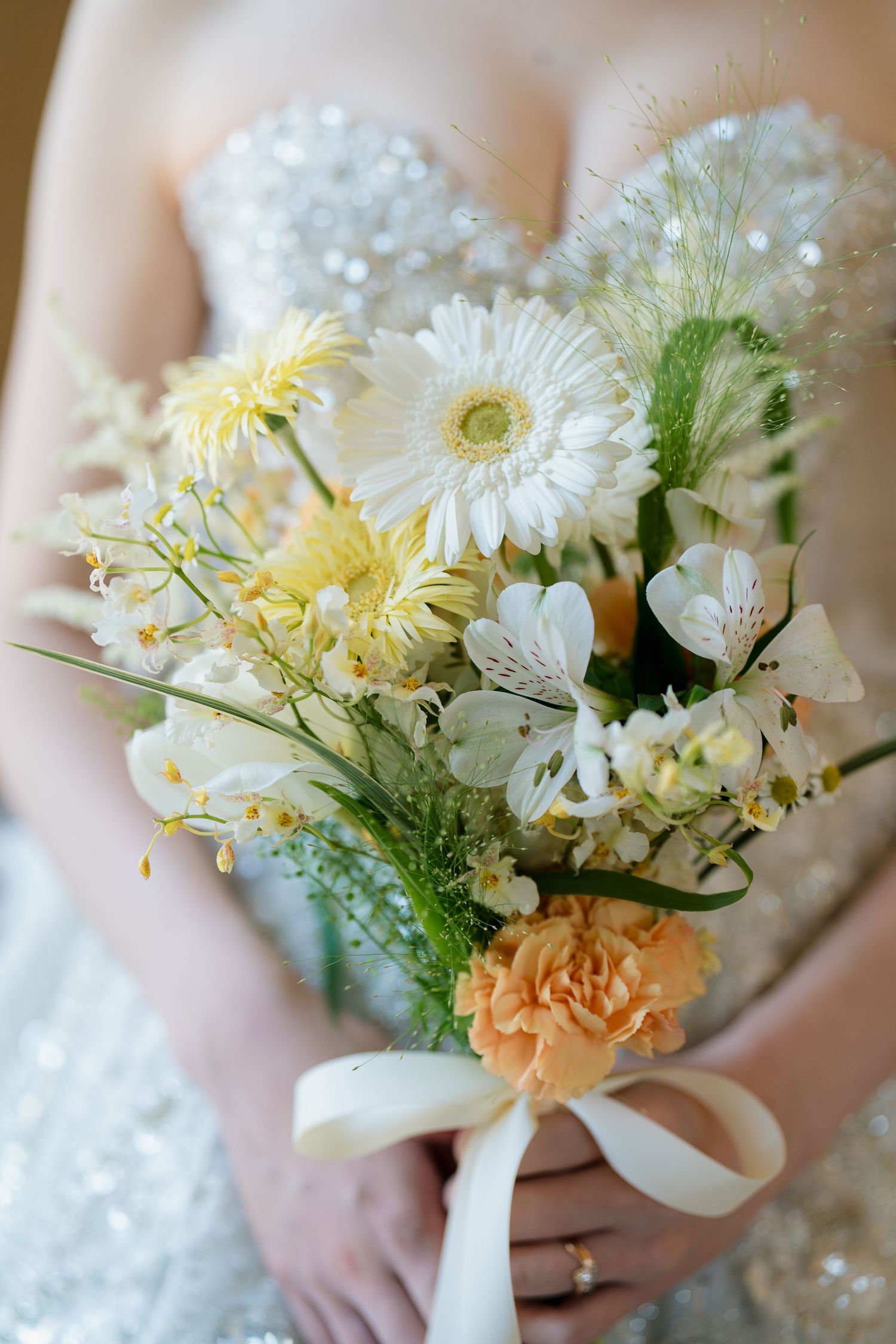 Bride holding a wedding bouquet of white gerbera daisies, yellow flowers, and an orange carnation tied with a white ribbon.