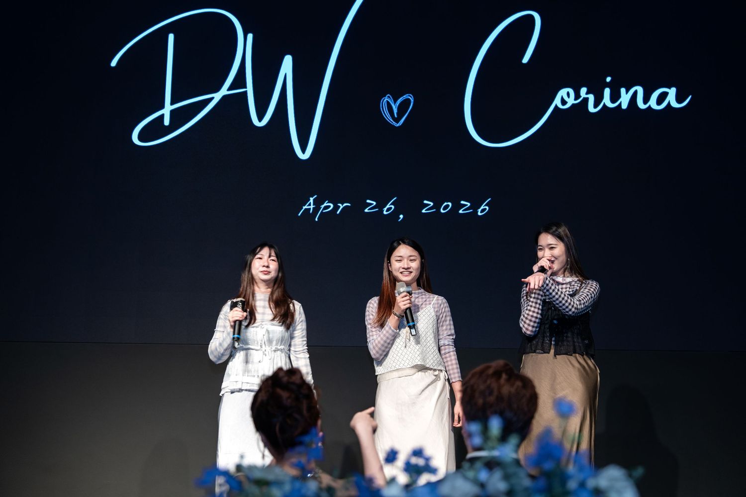 Three women speak at a wedding reception under a glowing sign reading DW and Corina, Apr 26, 2026.