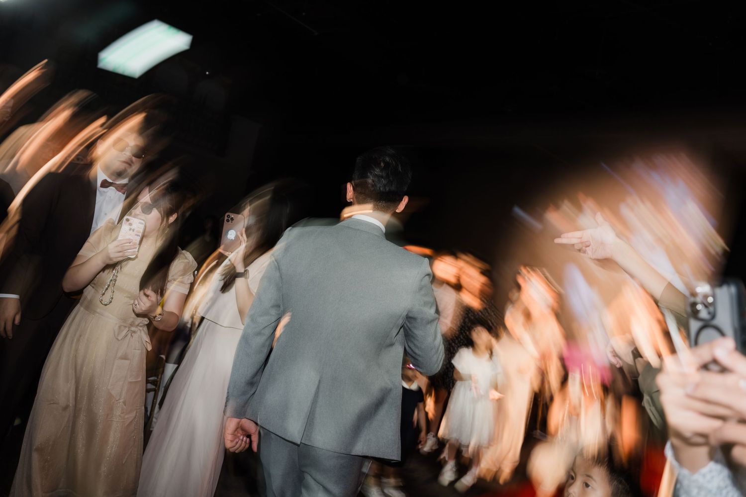 Motion-blurred party scene shows guests dancing at a dimly lit reception with someone in a gray suit in the center.
