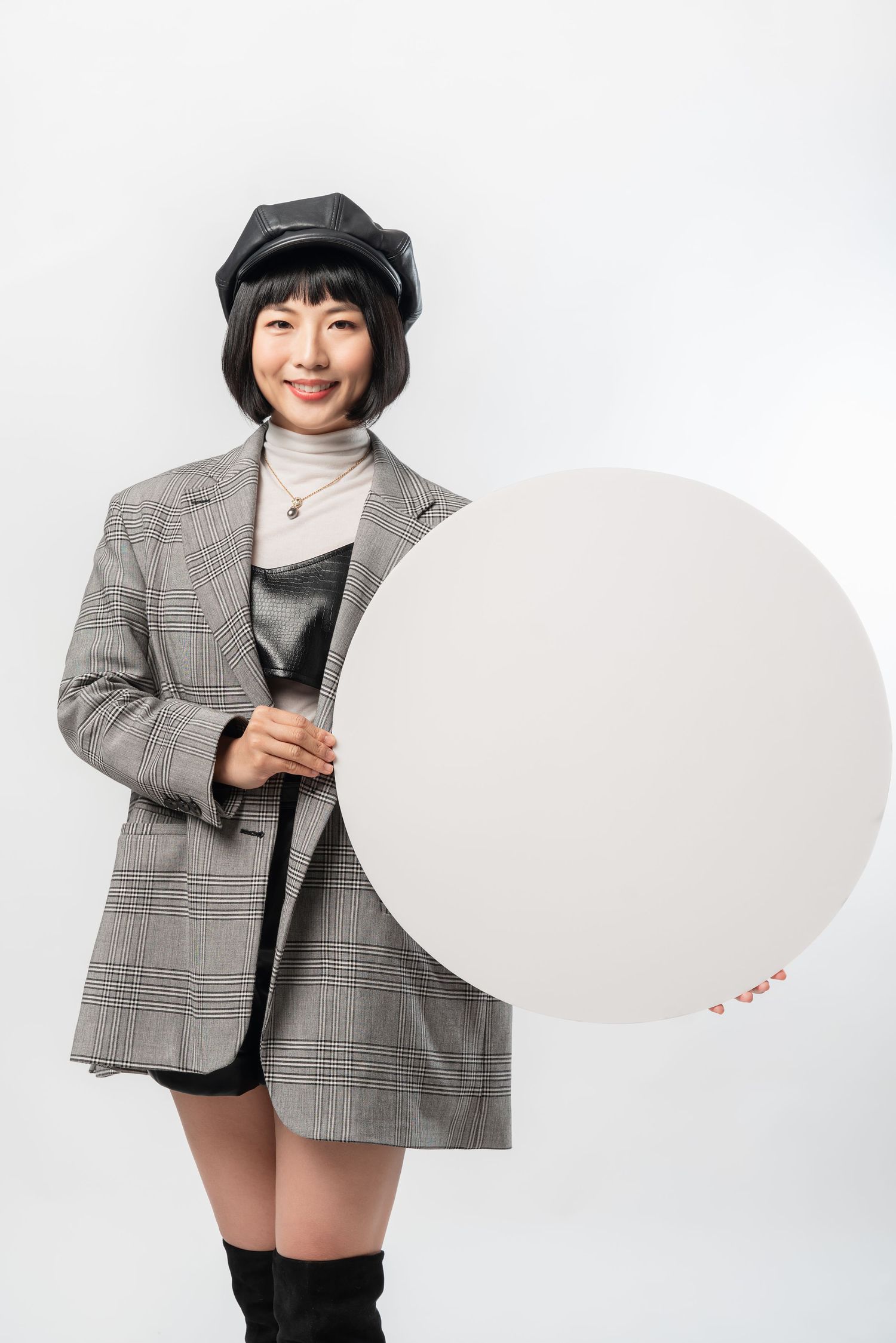 Stylish person in plaid blazer, black beret and boots holds blank white circular sign against white background.