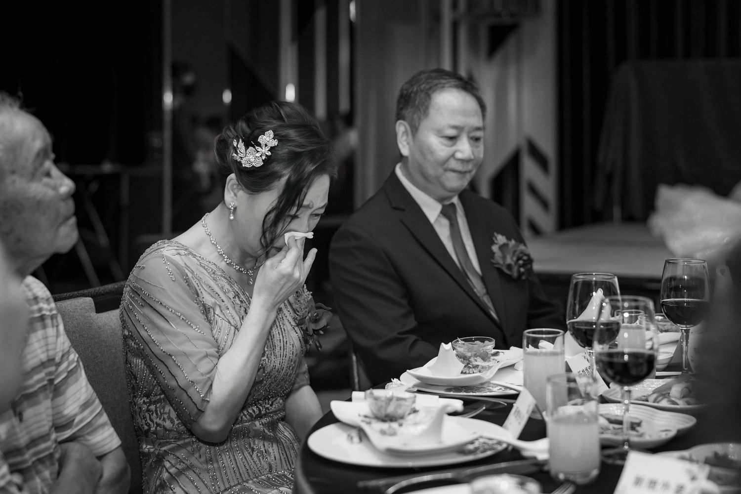A candid black and white photo of guests dining at an elegant restaurant table with wine glasses and formal attire.