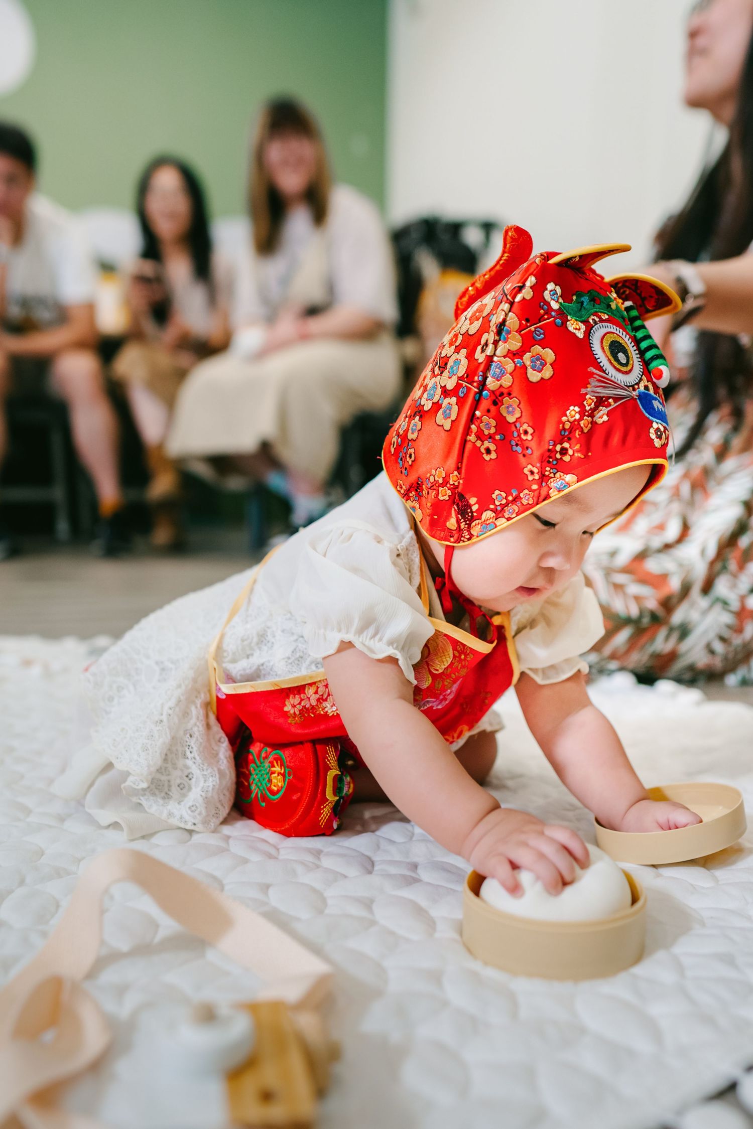 A baby wearing a red patterned outfit crawls on white fabric while reaching for a round wooden box.