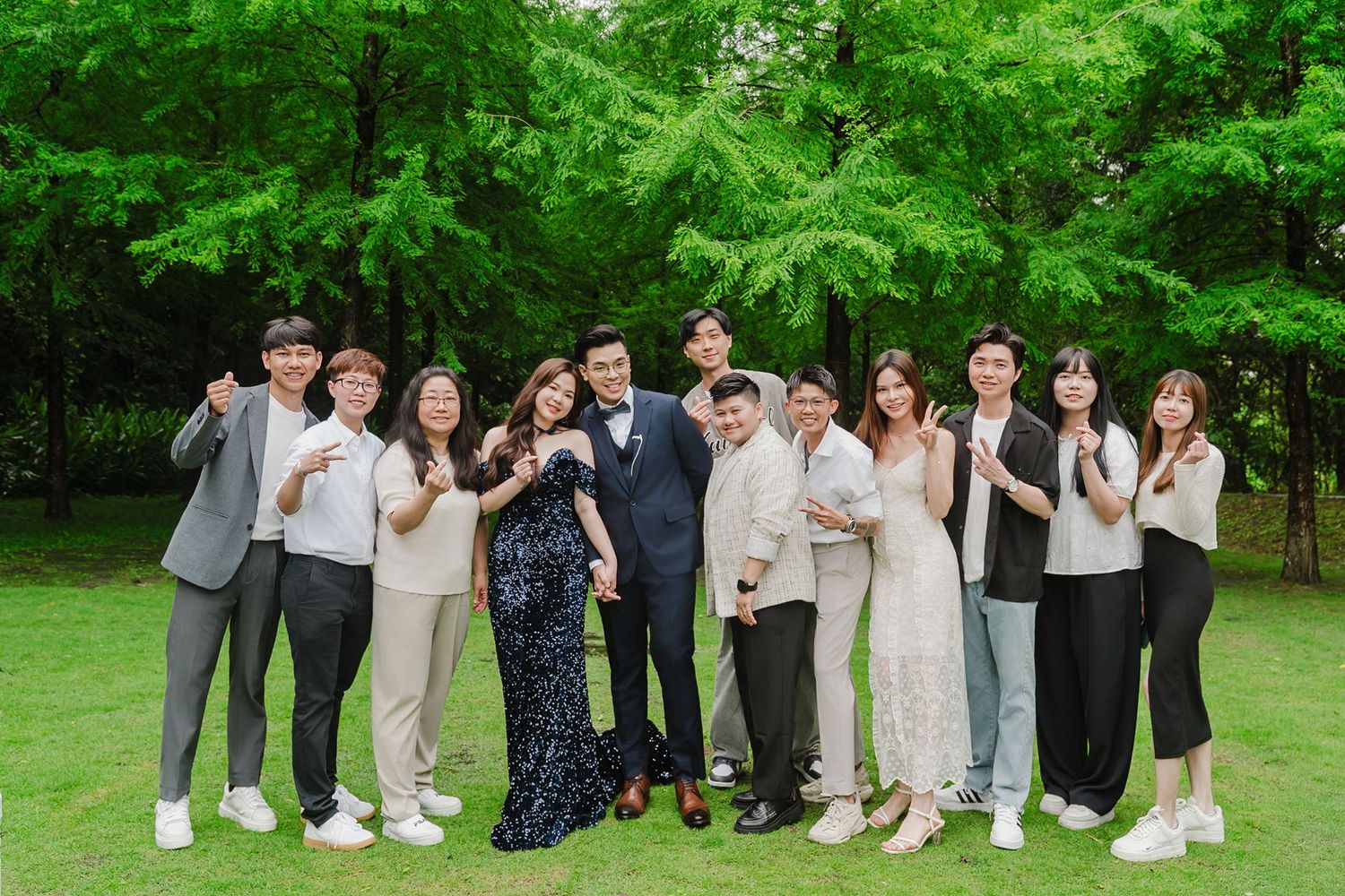 Wedding party group poses outdoors on grassy lawn in formal attire with lush green trees in background.