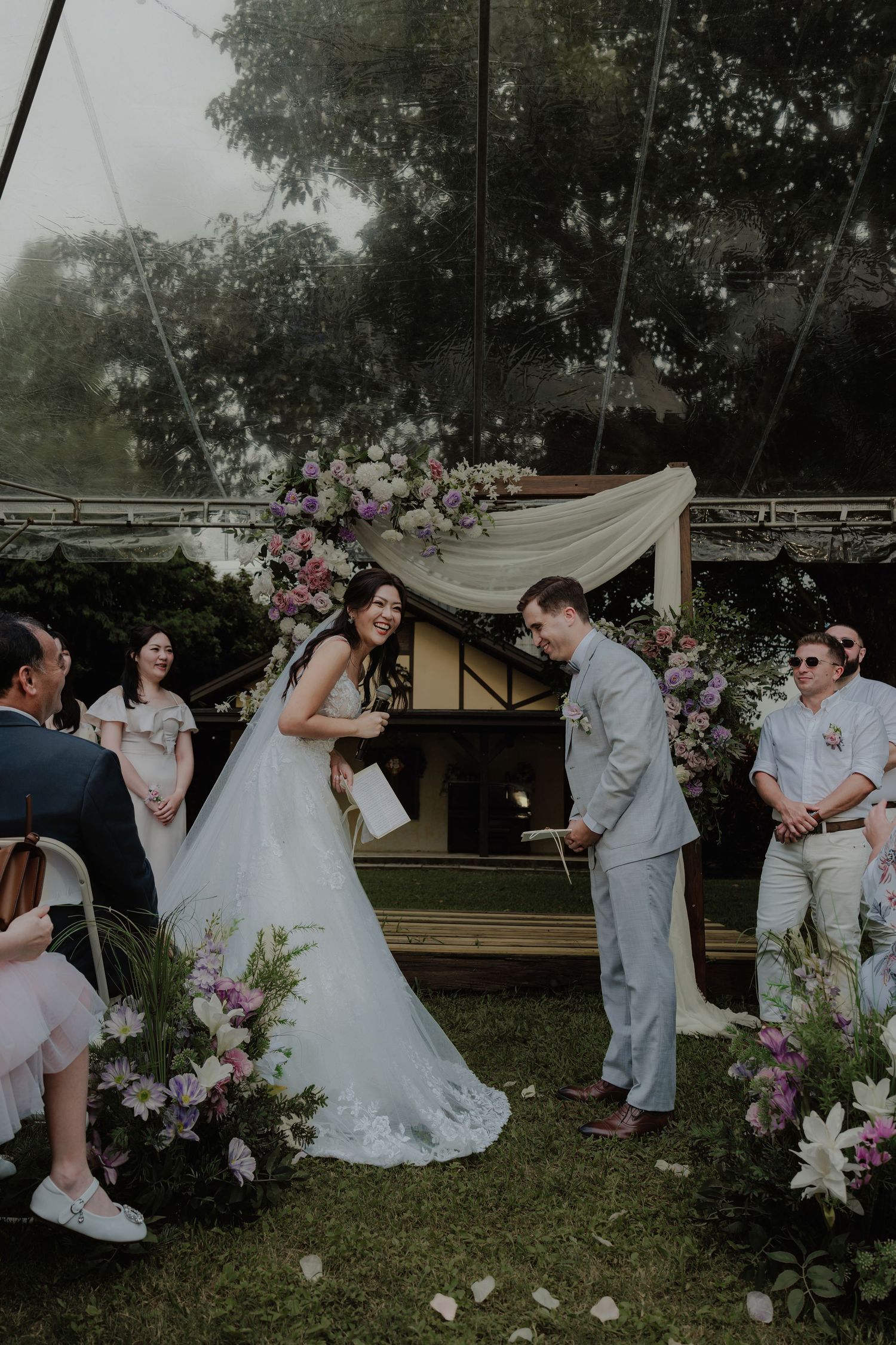 Bride and groom laughing together at outdoor wedding ceremony with purple floral arch and white draping.