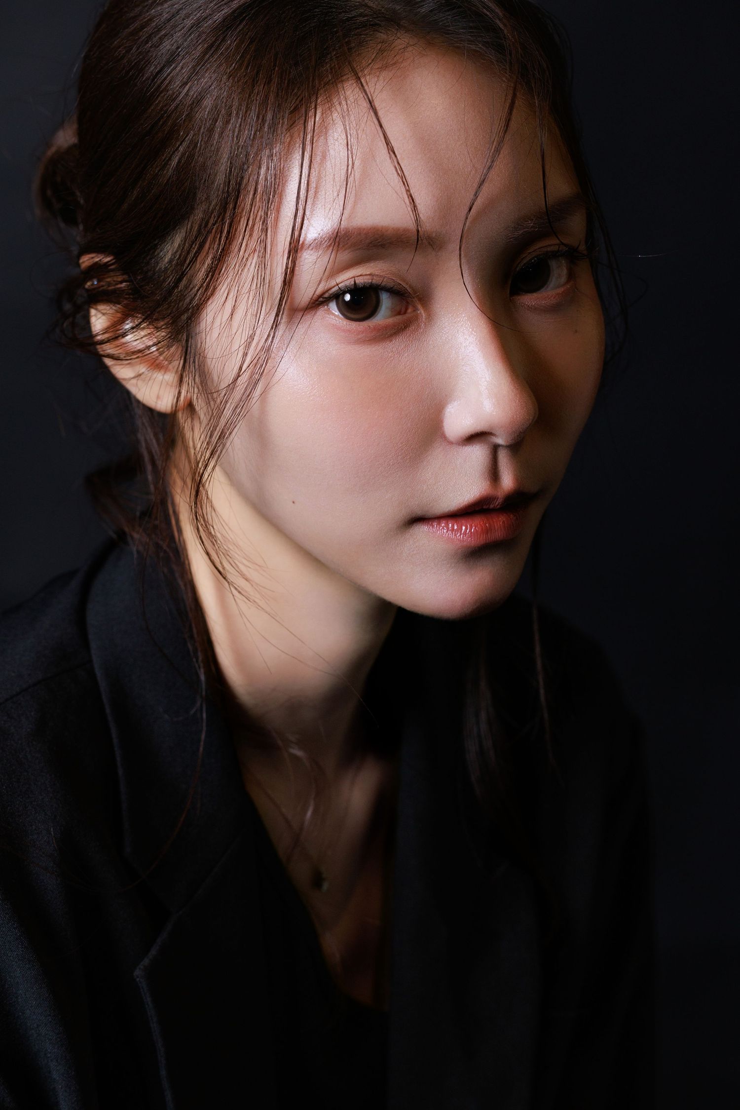 Close-up portrait of a young Asian woman with loose hair, wearing a black top, against a dark background.
