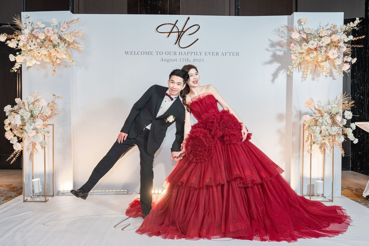 A couple poses playfully at their wedding with a dramatic red ballgown dress and black tuxedo in front of a decorated backdrop.