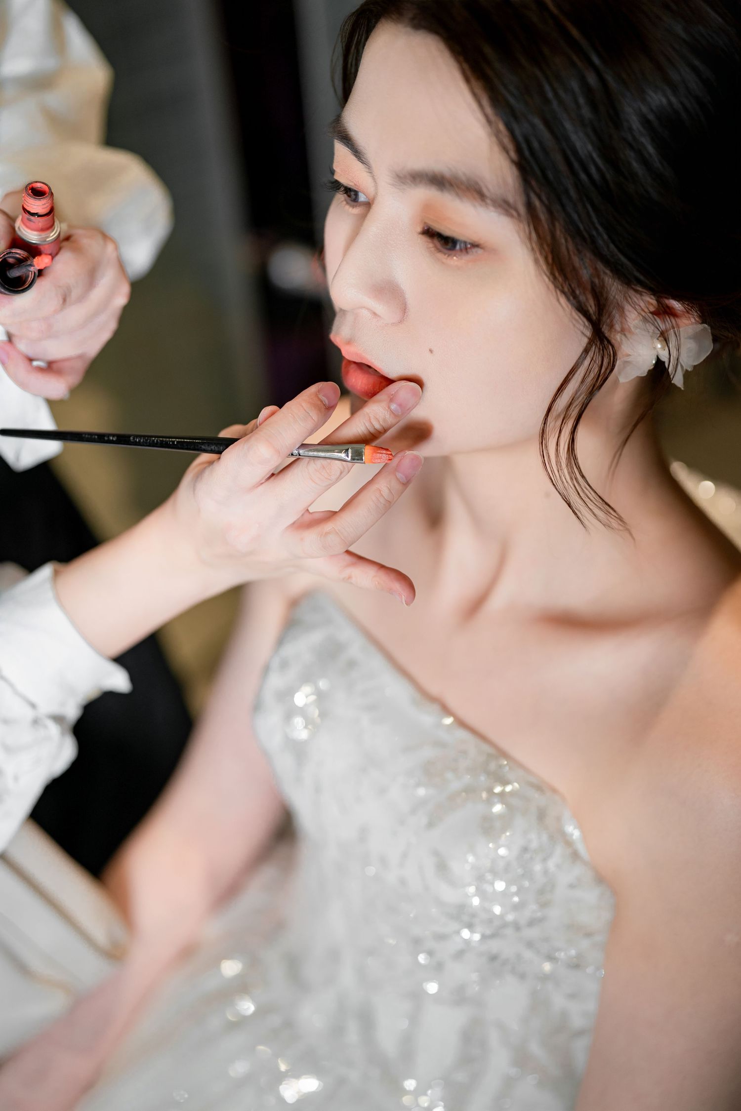 A makeup artist applies cosmetics to a person wearing a sparkly white dress during professional beauty preparation.
