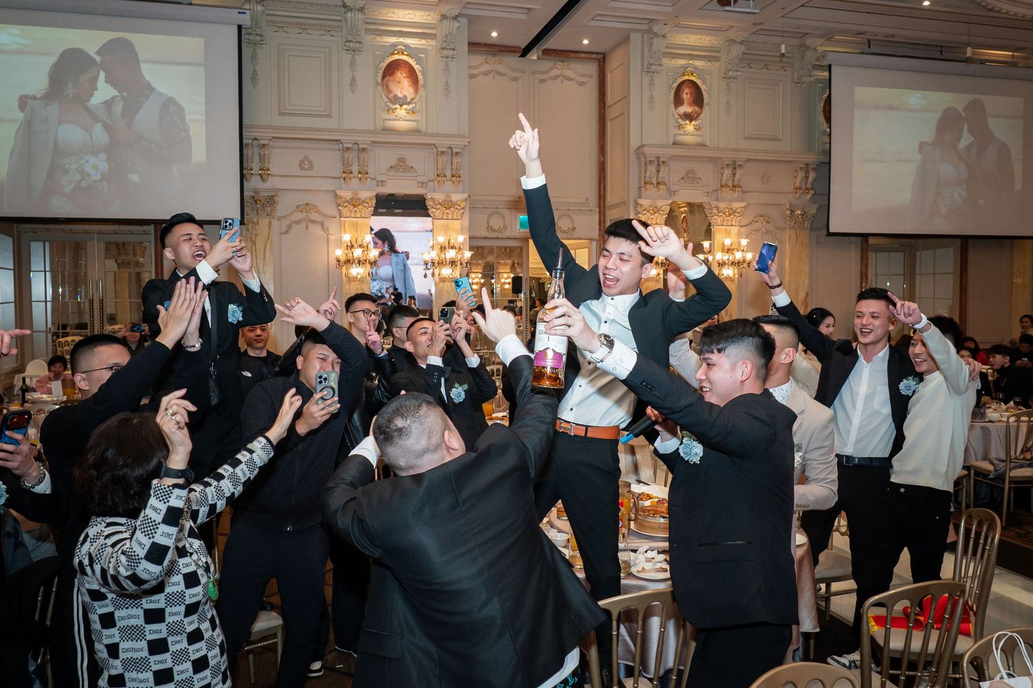 A group of wedding guests dance and celebrate together in a dimly lit reception venue with stone walls.