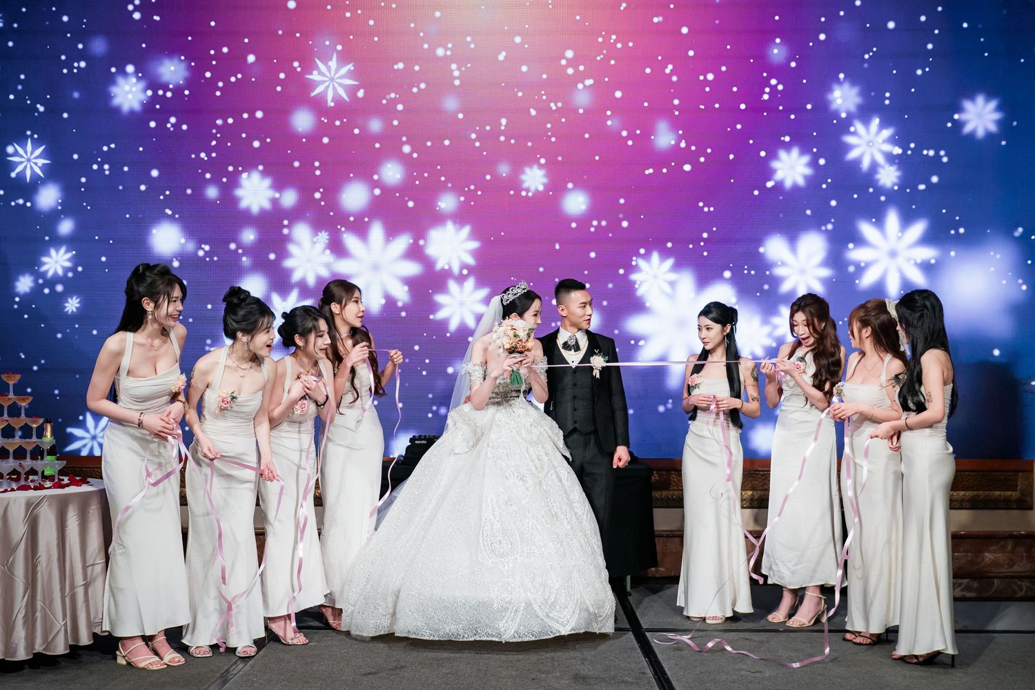 A group of bridesmaids in white dresses pose together against a magical winter backdrop with falling snowflakes.
