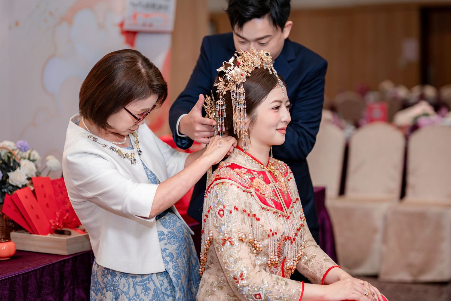 Two people help a bride in a red and gold embroidered Chinese wedding gown adjust her ornate phoenix headdress at a banquet venue.