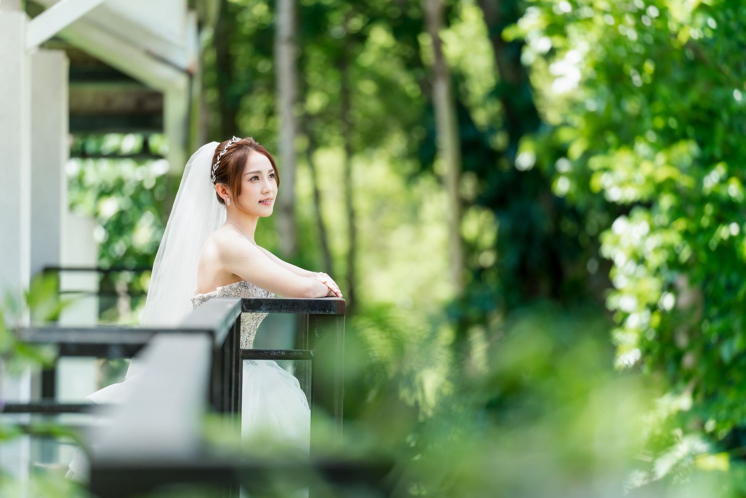 Bride in white ballgown leans thoughtfully on outdoor balcony railing surrounded by greenery.