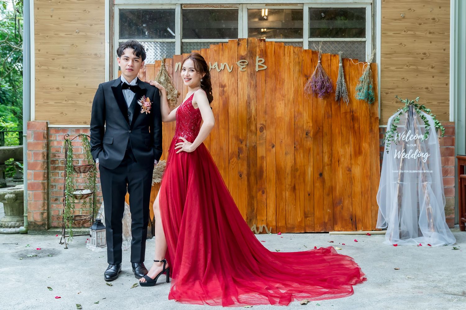 Elegant formal wear photo with red gown and black tuxedo against wooden wall with hanging decor.