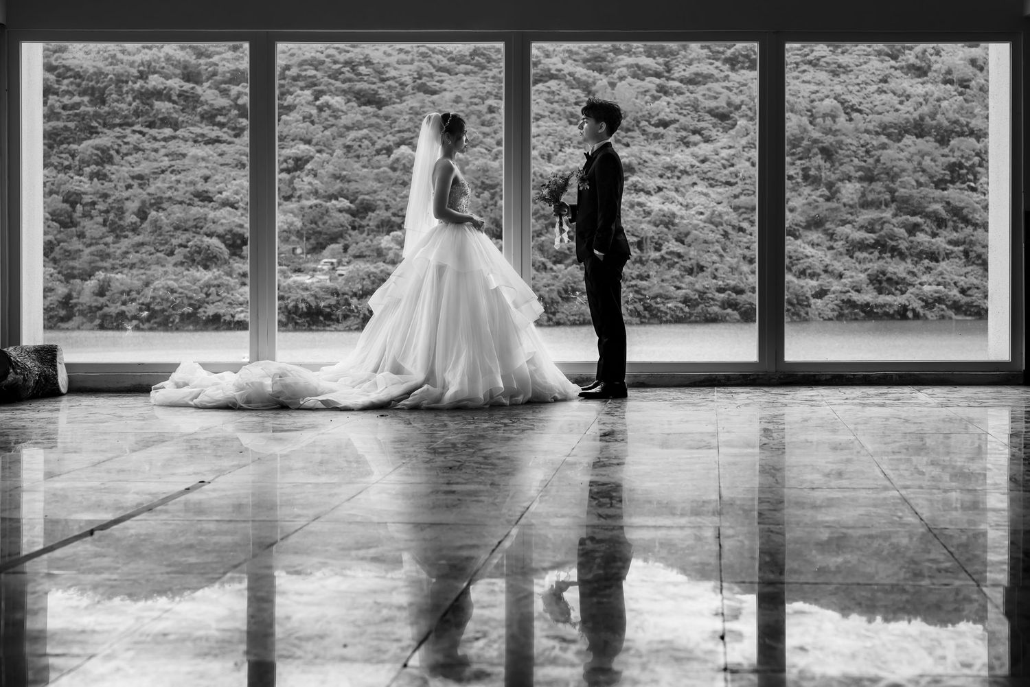 Silhouette of bride and groom standing by large windows with reflection on polished floor.
