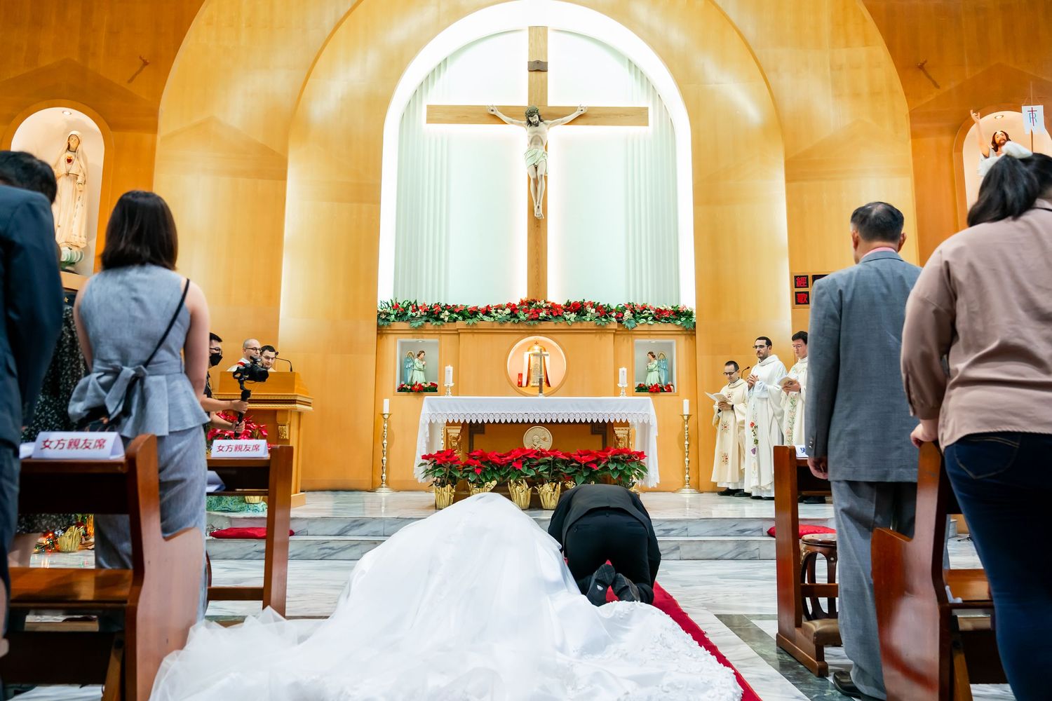 A wedding ceremony takes place in a Catholic church with a large crucifix and floral decorations on the altar.