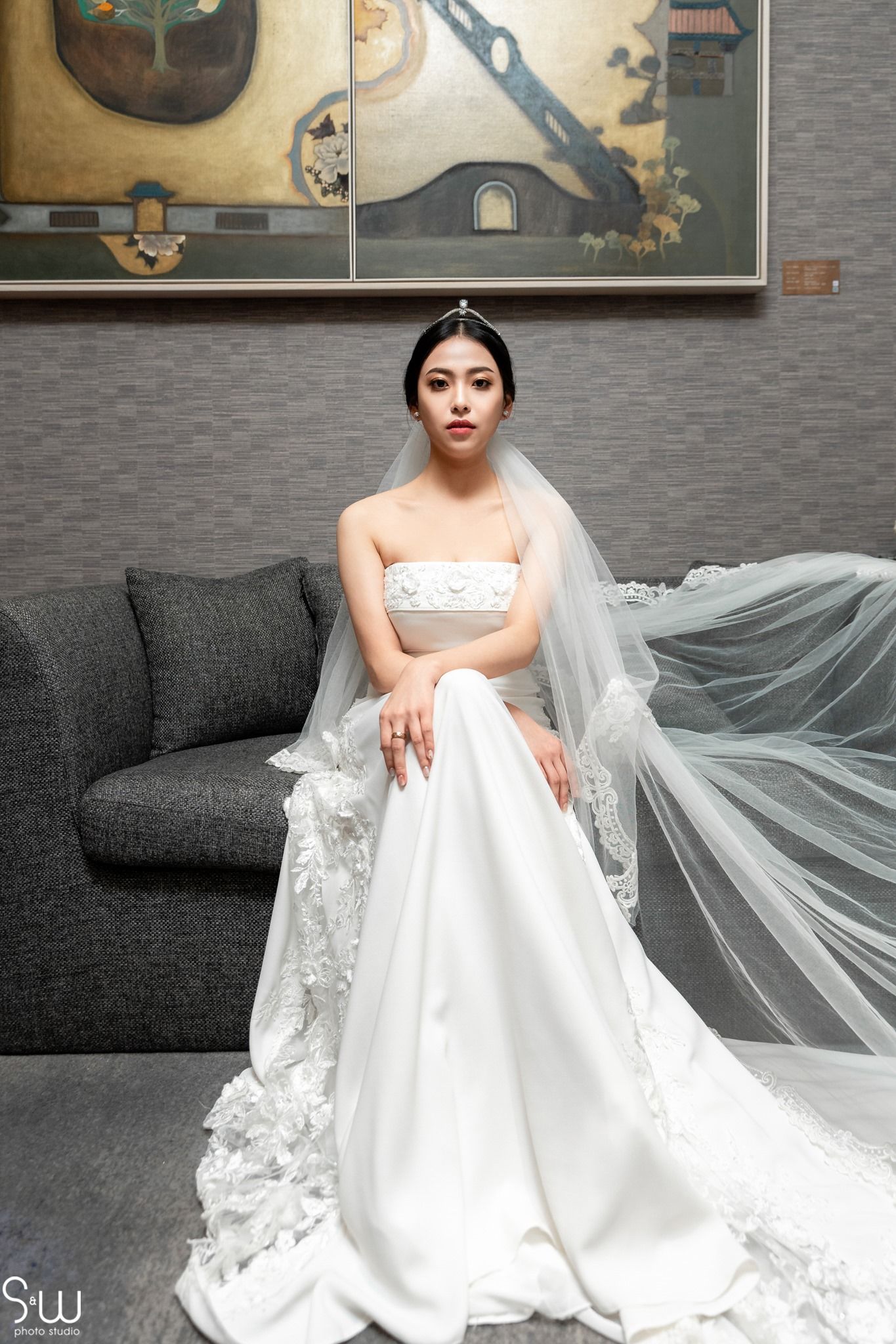 A bride in a stunning white lace wedding gown and veil sits elegantly on a gray couch against a textured wall.